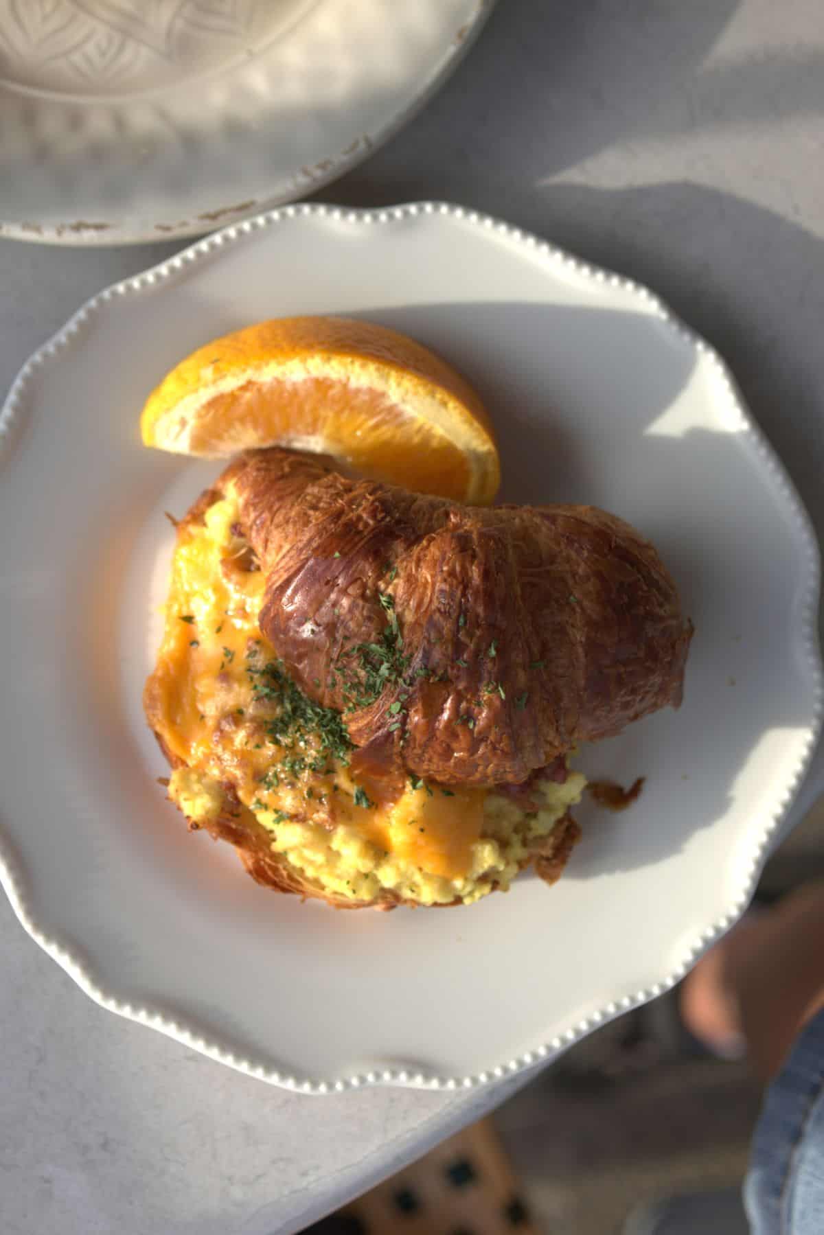 A croissant filled with scrambled eggs and cheese, garnished with herbs, served with an orange slice on a white plate.