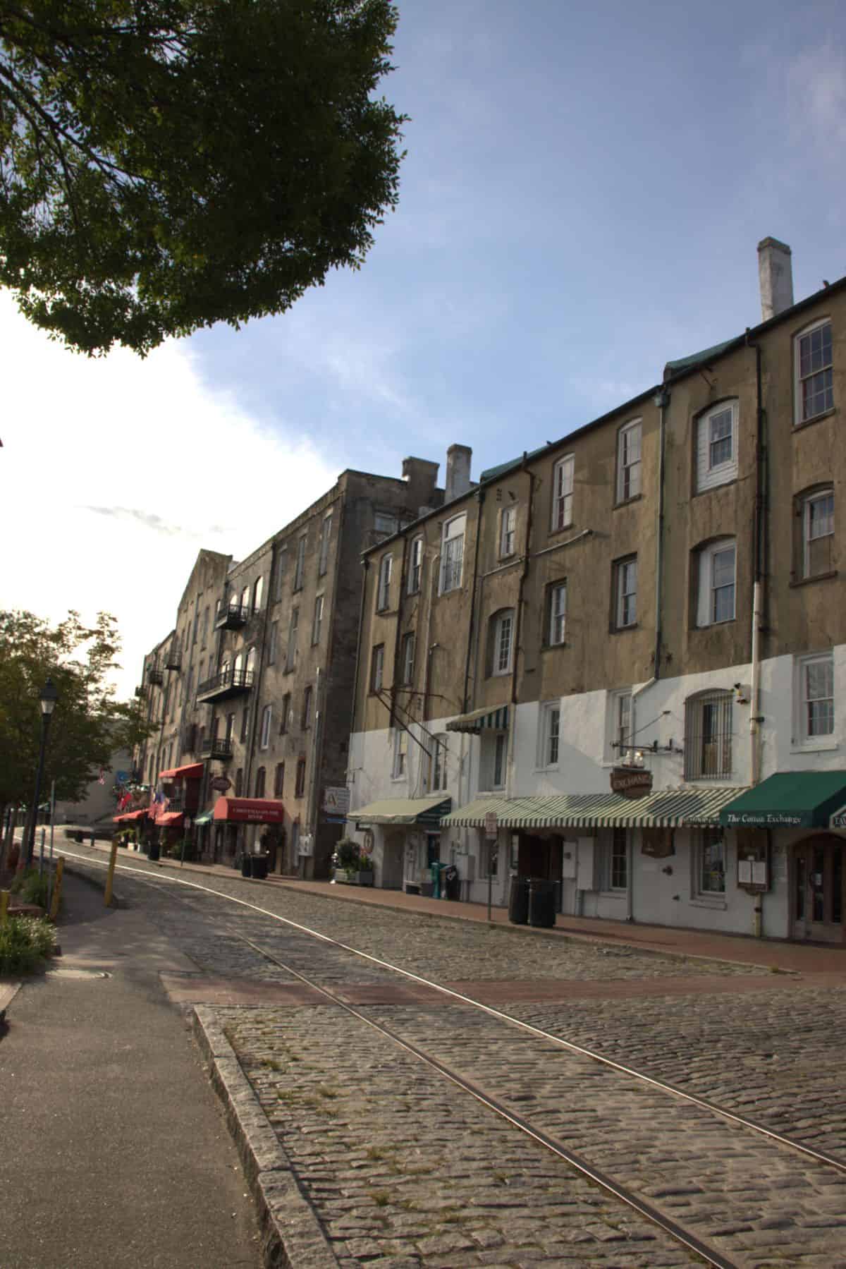 A row of historic multi-story buildings with awnings and storefronts lines a cobblestone street with tram tracks under a partly cloudy sky.