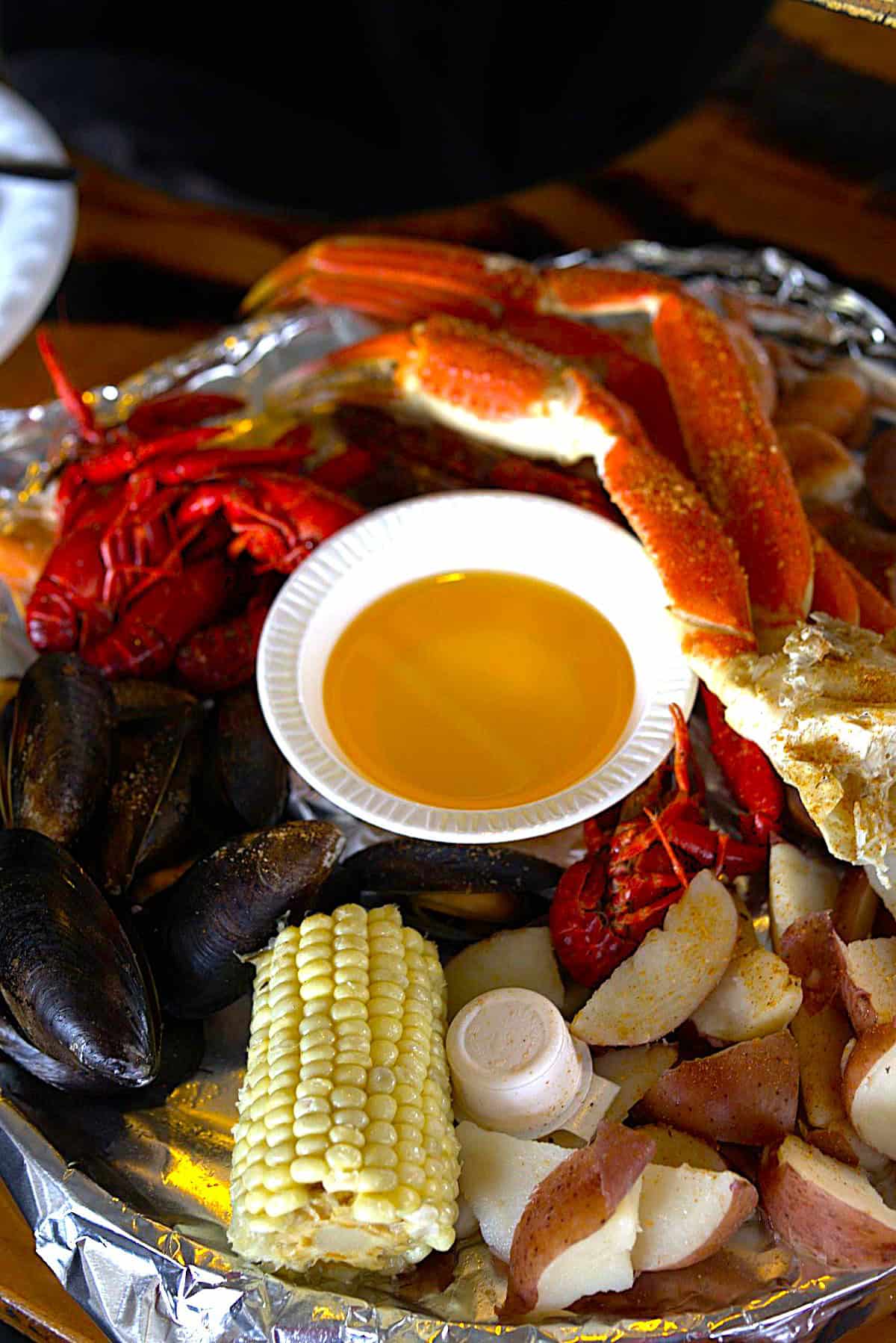 A seafood boil with crab legs, crawfish, mussels, potatoes, corn, and a cup of melted butter on a foil-lined tray.