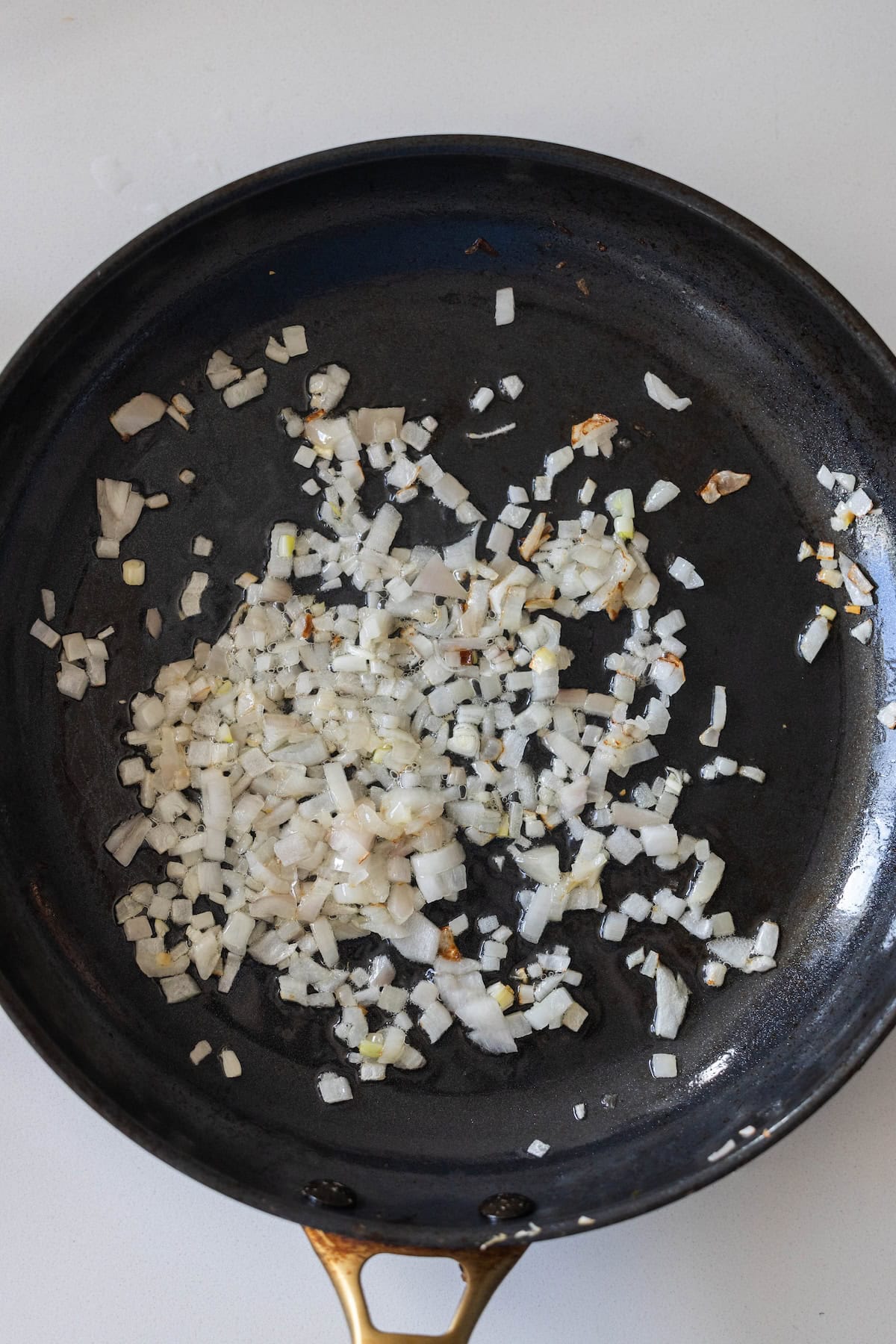 Chopped onions being sautéed in a black frying pan on a white surface, perfect as a savory base for Tuscan roasted white beans.
