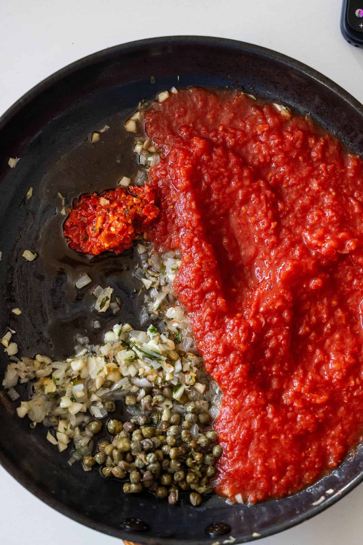 A frying pan with chopped onions and herbs, capers, tomato sauce, Tuscan Roasted White Beans, and a spoonful of red paste, all separated and uncooked.