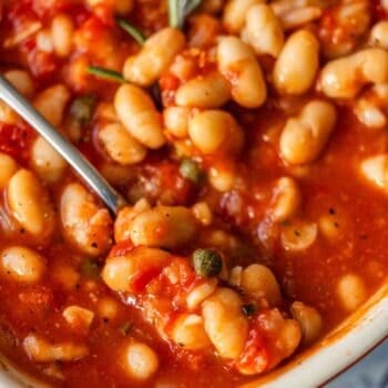 Close-up of Tuscan roasted white beans in a tomato-based sauce with herbs, served in a dish with a spoon.