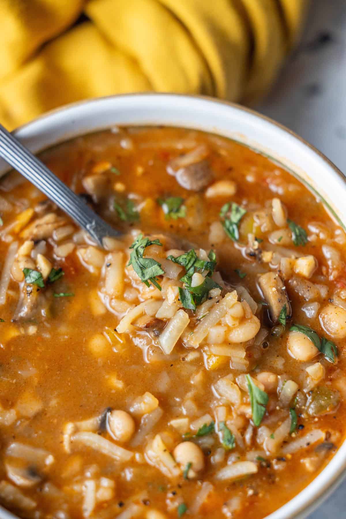 A bowl of hearty vegan soup with rice, chickpeas, mushrooms, and herbs, served with a spoon and set against a yellow cloth in the background.
