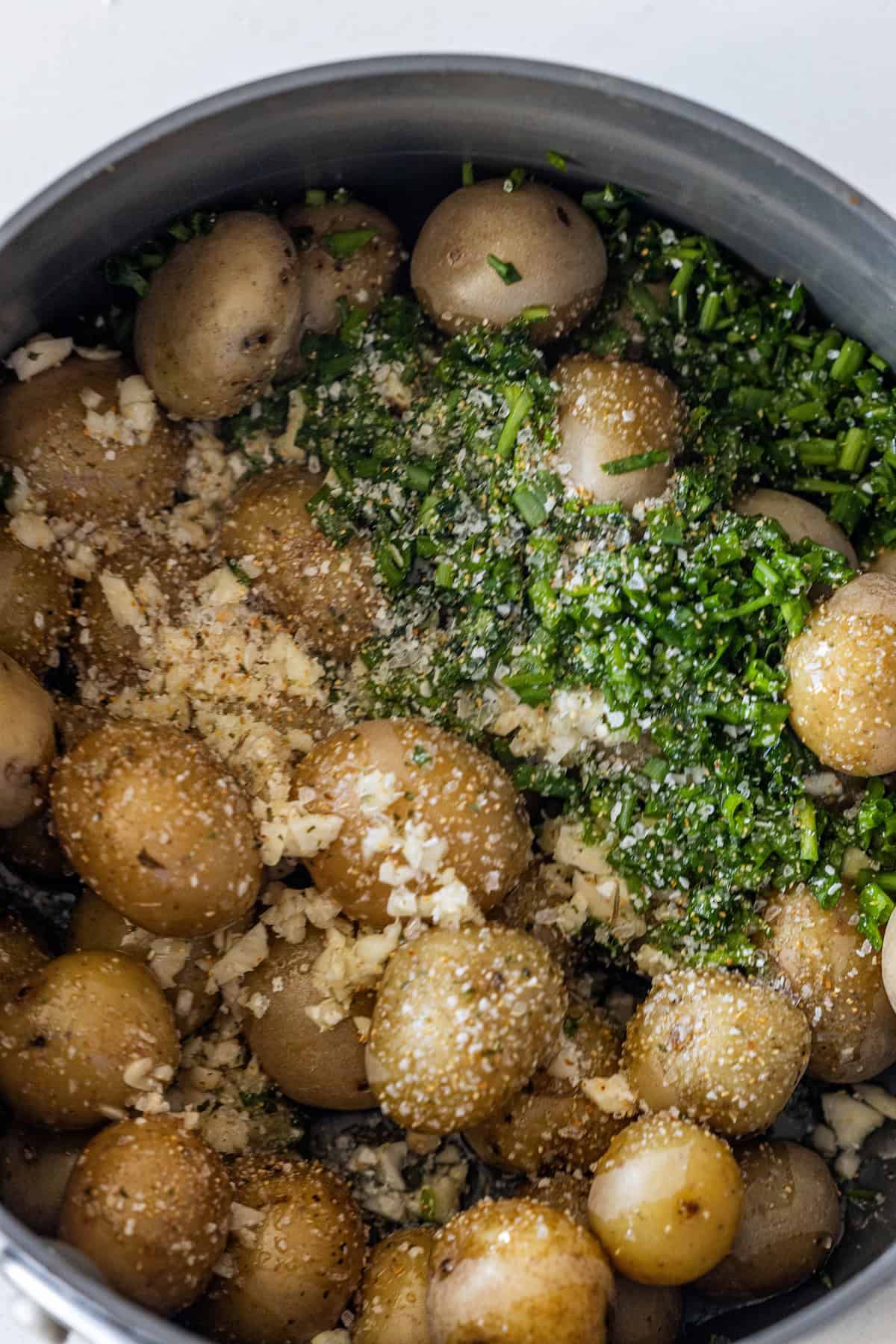 A pot filled with baby potatoes, chopped herbs, minced garlic, grated cheese, and seasonings is ready to be mixed or cooked into delicious garlic and chive potatoes.