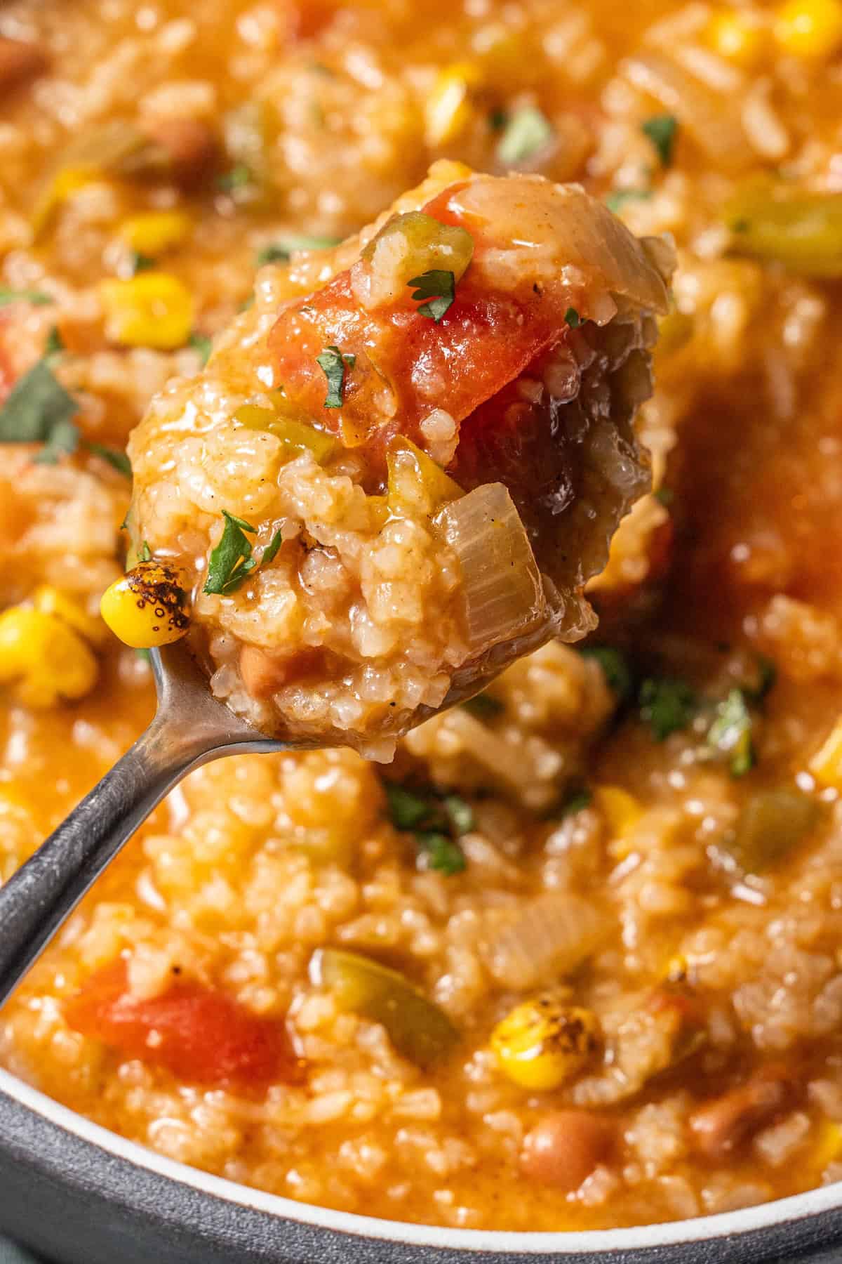 A close-up of a spoonful of thick vegan soup with corn, tomatoes, rice, and herbs held above a pot of hearty rice soup.