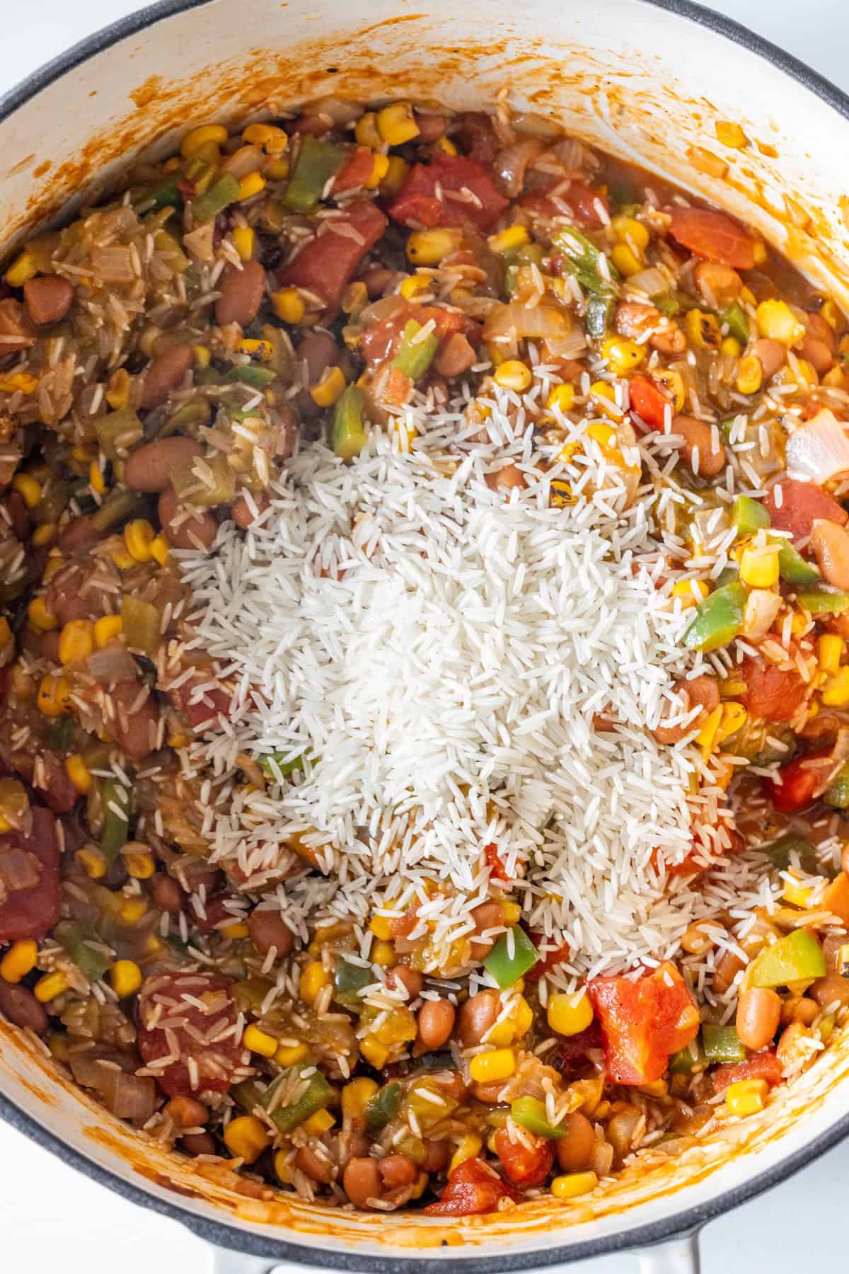 A pot filled with a mixture of charro beans, corn, tomatoes, peppers, and uncooked white rice on top, ready to be stirred into a hearty vegan soup.