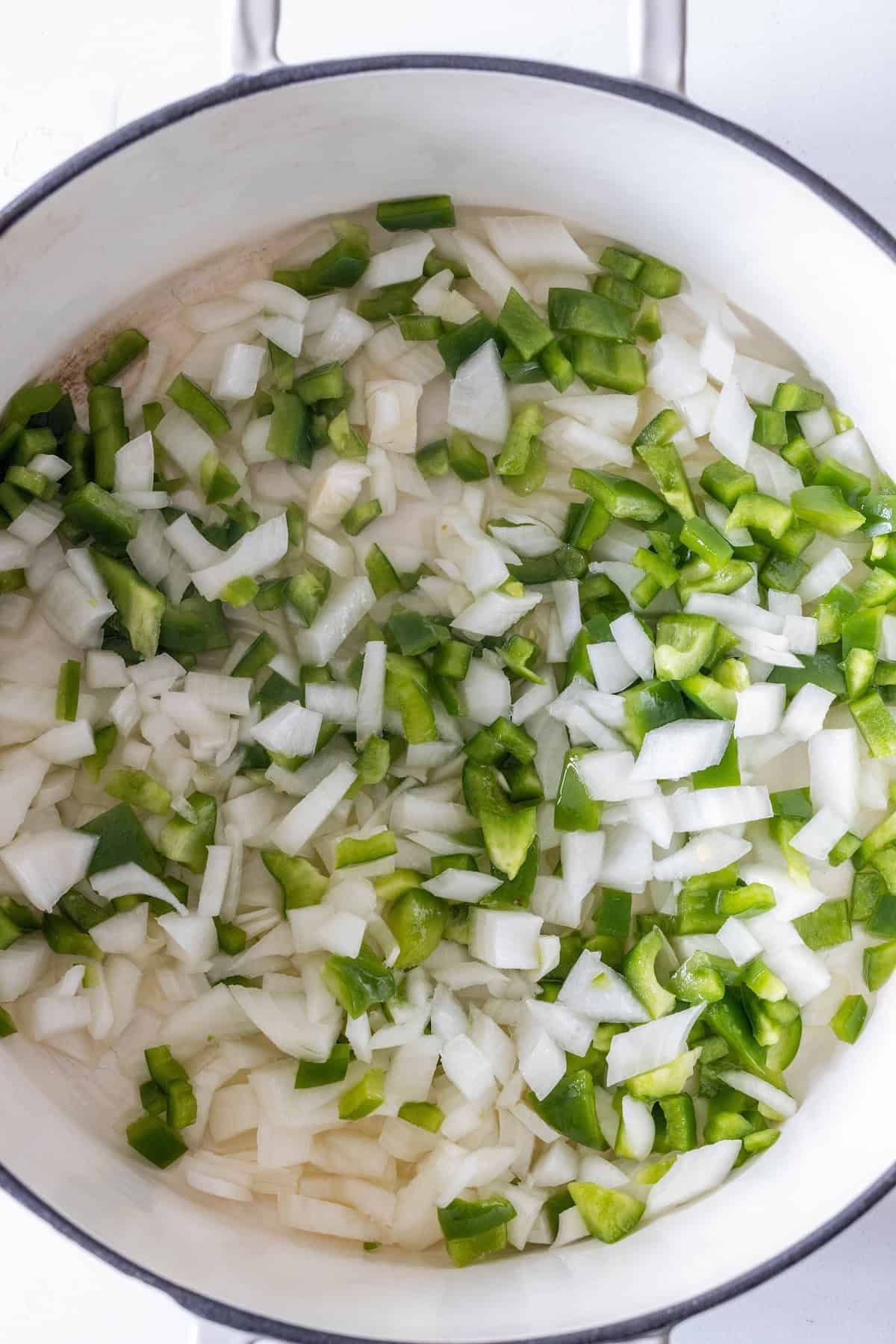 Chopped white onions and green bell peppers in a white pot, ready to be cooked for a flavorful vegan rice soup.