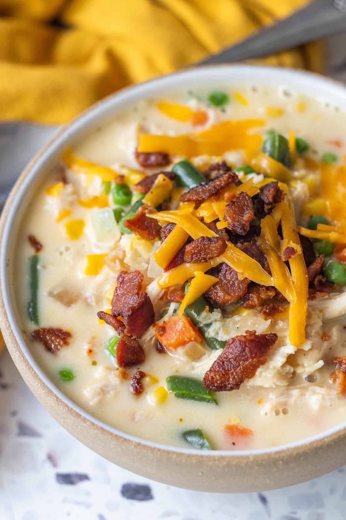 A bowl of creamy Cheesy Corn Chowder with green beans, carrots, corn, shredded cheddar cheese, and crispy bacon pieces on top.