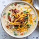 A bowl of creamy corn chowder with mixed vegetables, shredded cheddar cheese, and crispy bacon pieces on top, next to a spoon and a yellow napkin.