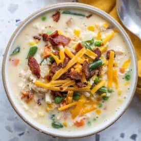 A bowl of creamy corn chowder with mixed vegetables, shredded cheddar cheese, and crispy bacon pieces on top, next to a spoon and a yellow napkin.