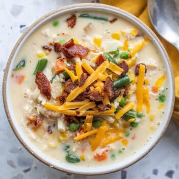 A bowl of creamy corn chowder topped with shredded cheddar cheese, crispy bacon, and filled with mixed vegetables like peas, green beans, carrots, and corn. A spoon and napkin are nearby.