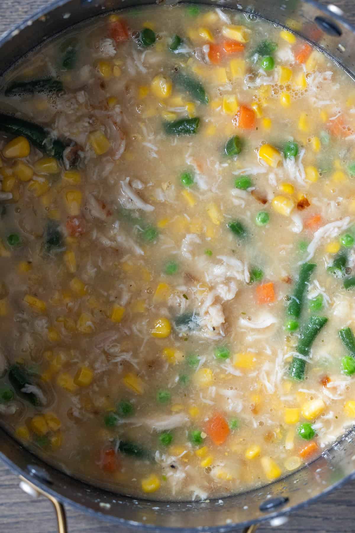 A close-up of a pot filled with thick cheesy corn chowder featuring bacon, green beans, peas, carrots, and tender shredded chicken.