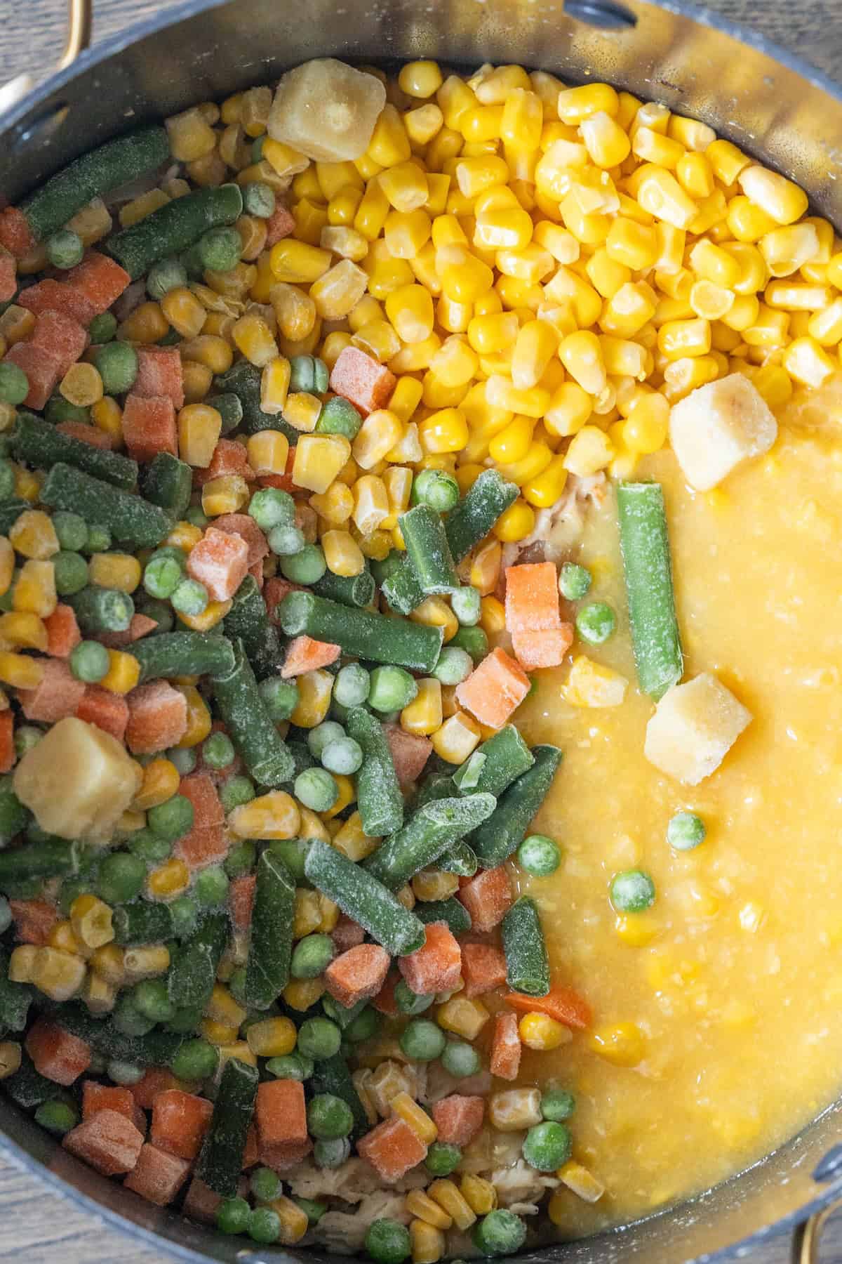 A pot contains frozen mixed vegetables, frozen corn, and a thick yellow liquid, partially blended together for a hearty Cheesy Corn Chowder.