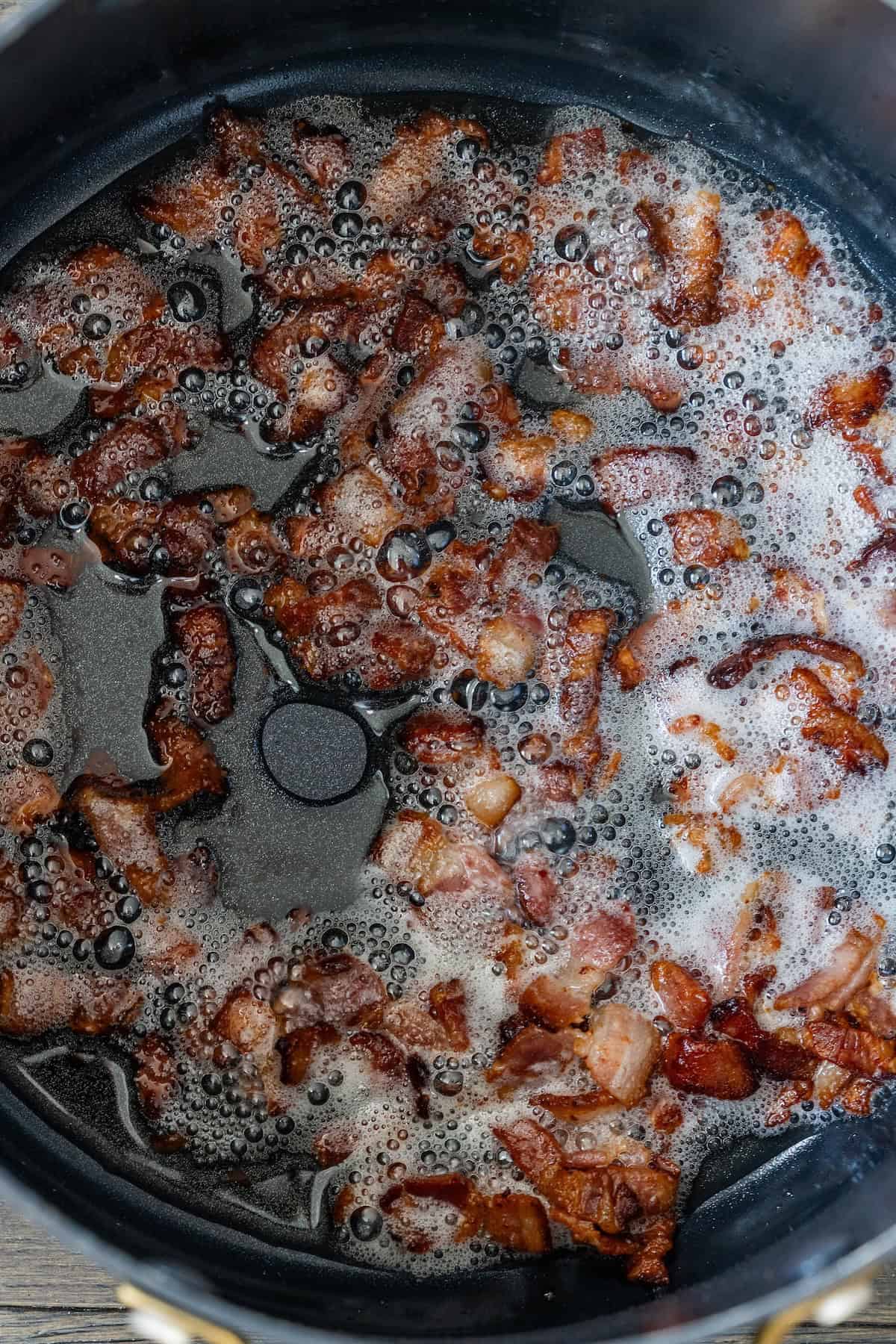 Crispy bacon pieces cooking in a black pan with bubbling grease and foam&mdash;perfect for topping cheesy corn chowder.