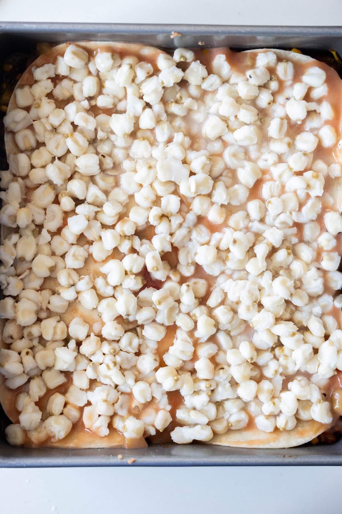 A baking pan filled with Vegan Enchilada Pie, featuring a layered dish topped with white hominy kernels and a light orange sauce.