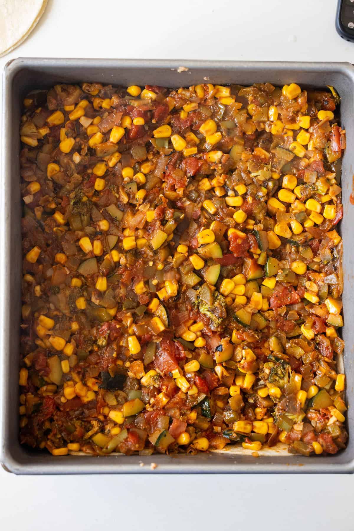 A square baking pan filled with a cooked mixture of corn, tomatoes, zucchini, and other vegetables in a tomato-based sauce creates a delicious Vegan Enchilada Pie.