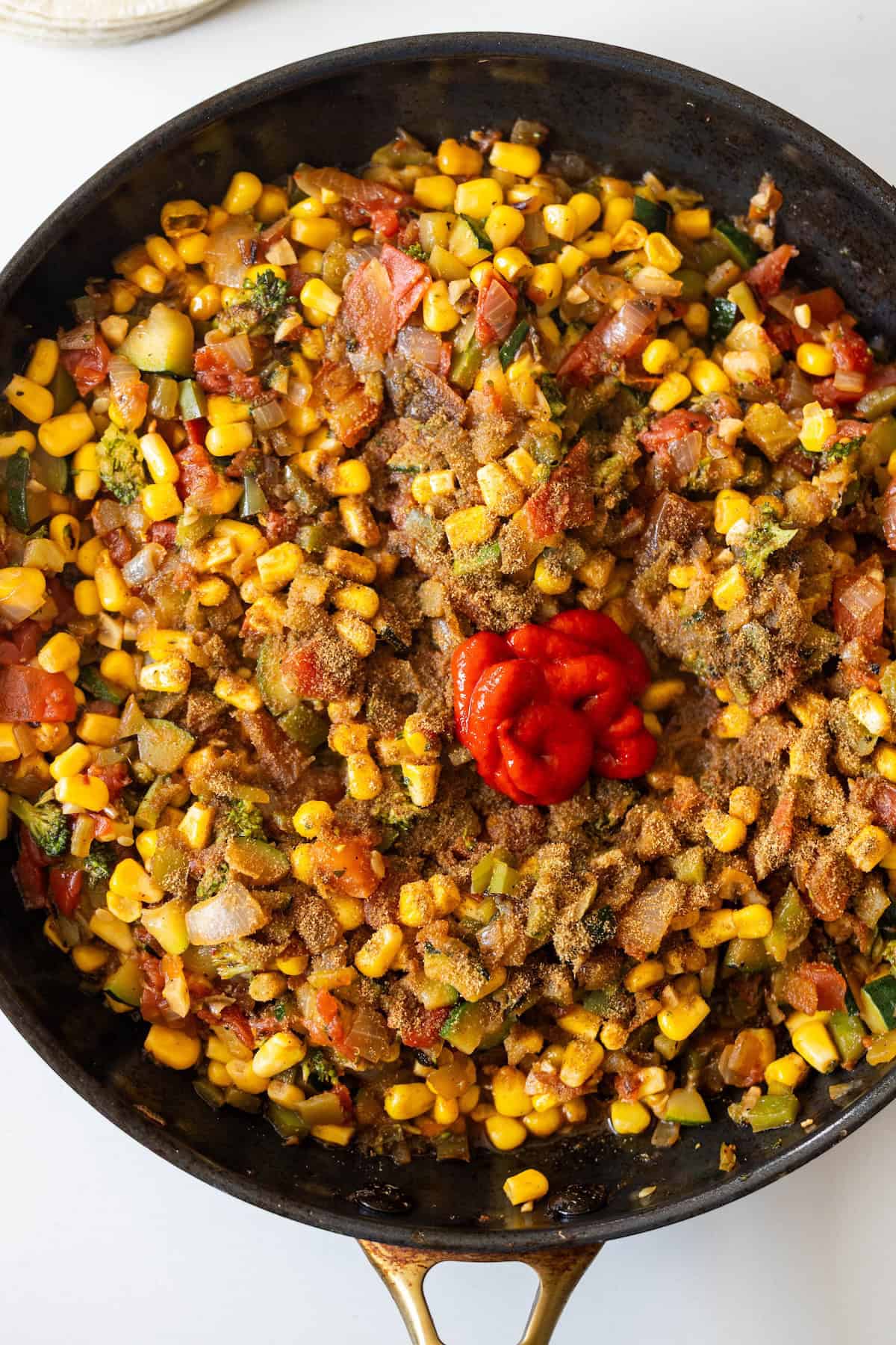 A skillet filled with sautéed corn, diced tomatoes, onions, and spices, reminiscent of a Vegan Enchilada Pie, with a dollop of tomato paste or ketchup in the center.