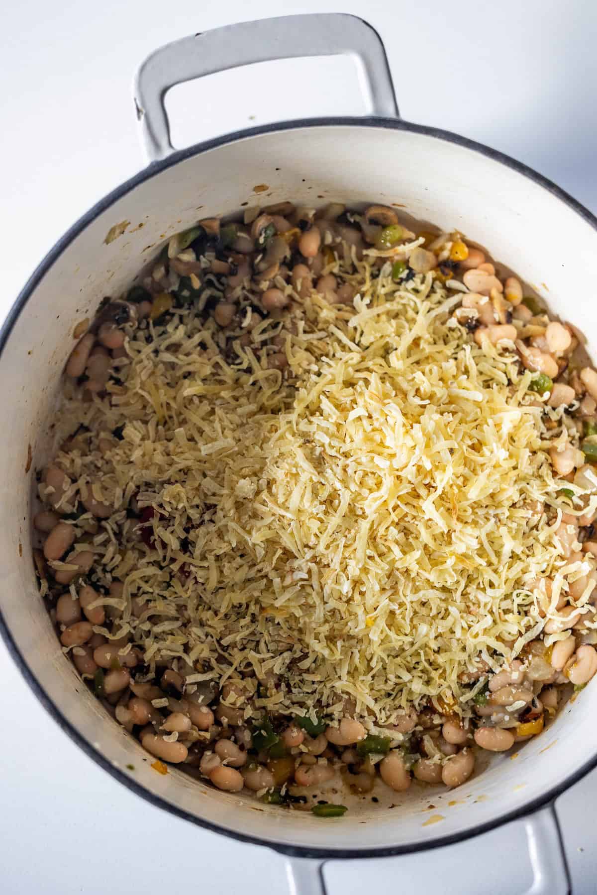 A white pot filled with cooked white beans, vegetables, and a heap of shredded cheese on top, ready to be mixed&mdash;a hearty vegan soup perfect for chilly days.