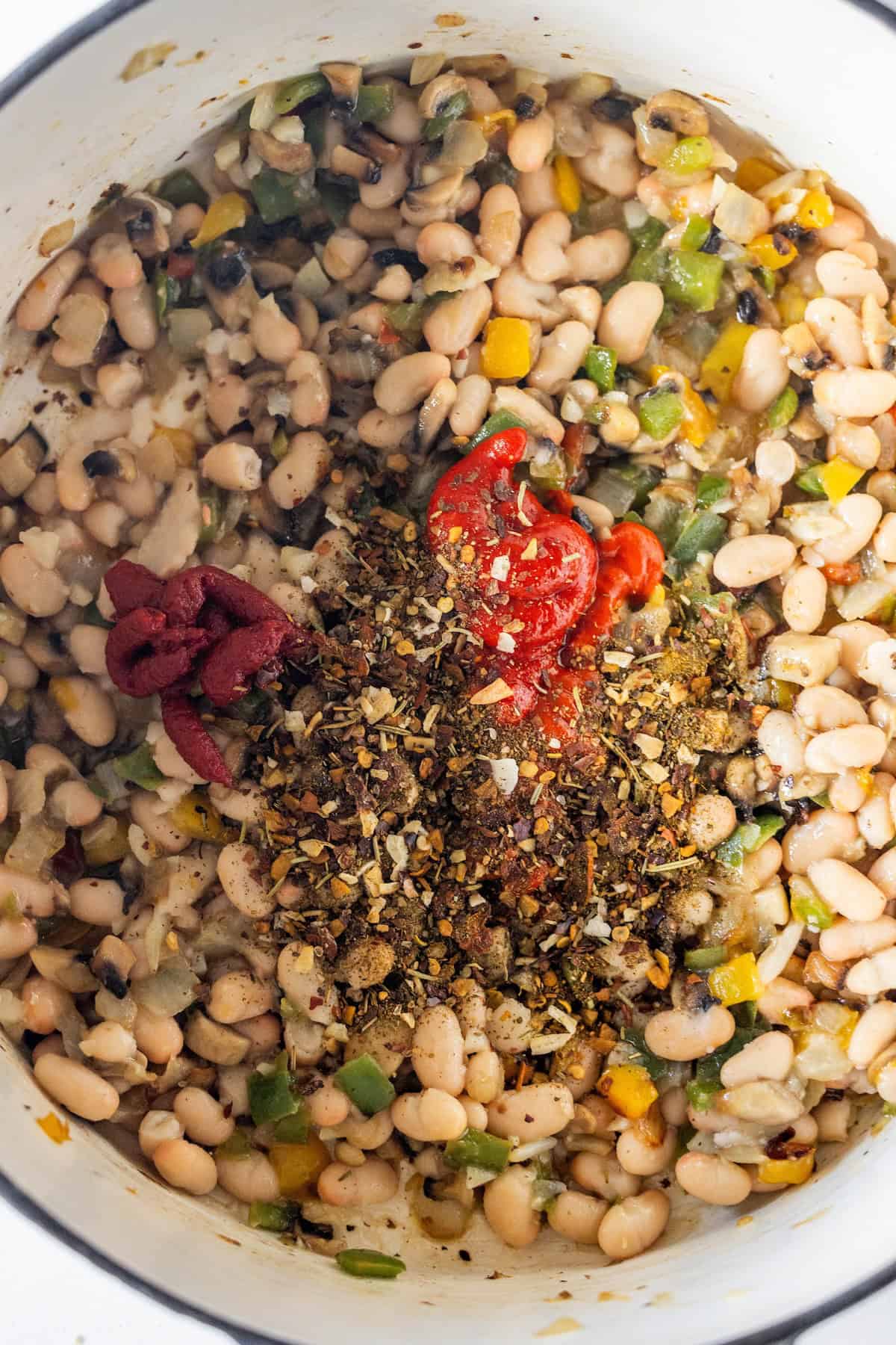 A pot of vegan soup with cooked white beans, chopped green and yellow vegetables, tomato paste, and a sprinkle of dried herbs and spices on top, not yet stirred.