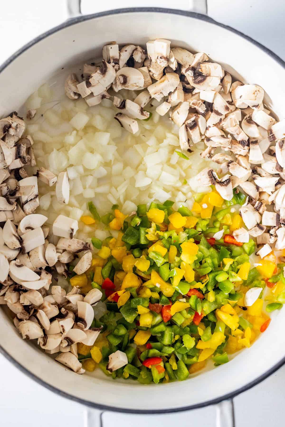 Chopped onions, mushrooms, and mixed bell peppers are arranged in sections inside a white pot, ready to be cooked for a delicious vegan soup.
