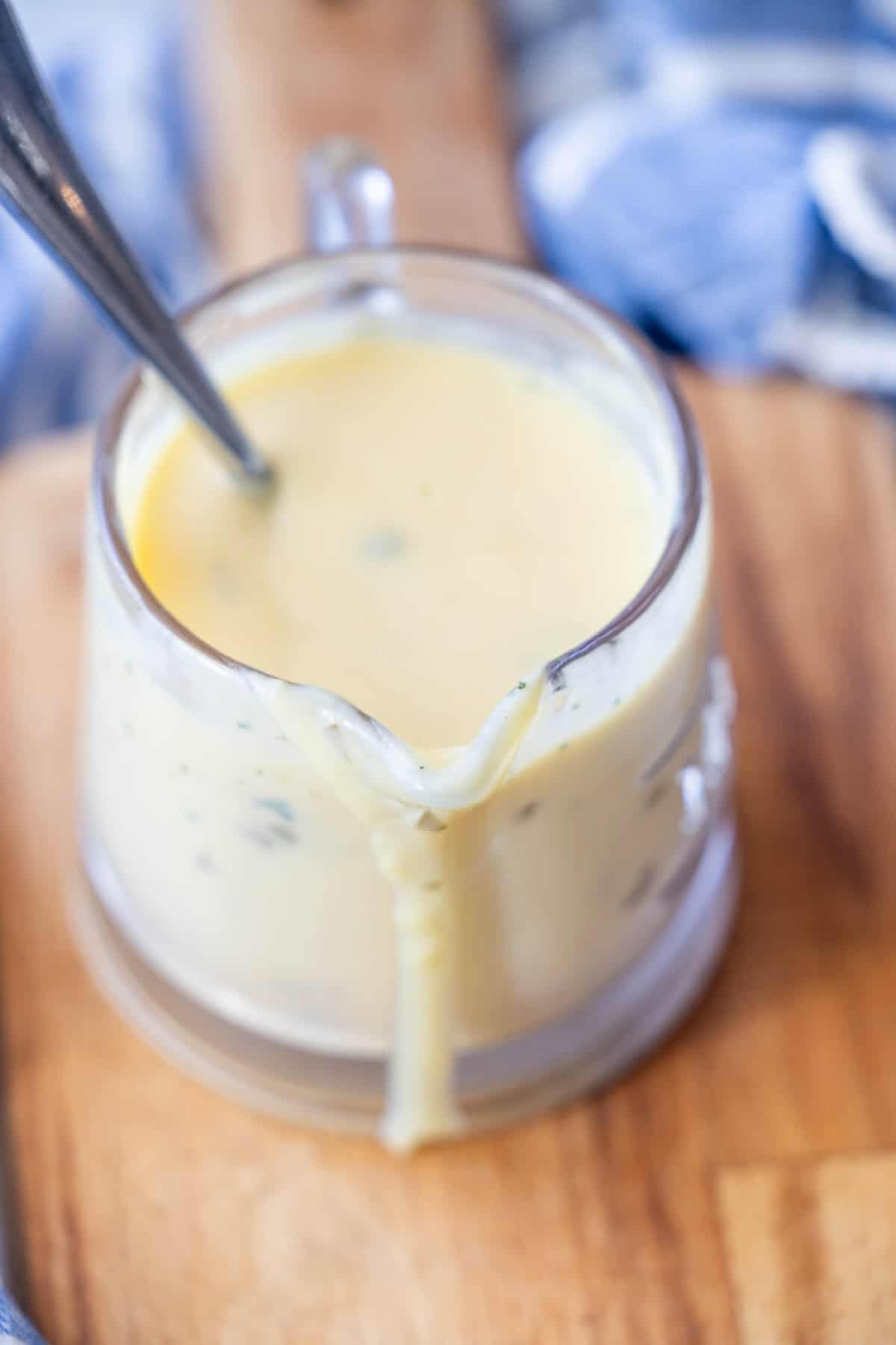 A glass measuring cup filled with creamy yellow avgolemono sauce, with a spoon inside and sauce dripping down the spout, sits on a wooden surface.