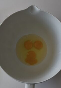 Three raw eggs cracked in a white mixing bowl, their yolks grouped together at the center&mdash;perfect beginnings for preparing silky Avgolemono Sauce.