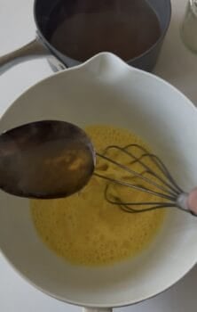 A metal spoon pours liquid into a bowl of beaten eggs being whisked, as Avgolemono sauce&mdash;a classic Greek sauce&mdash;comes together, with a saucepan and jar in the background.