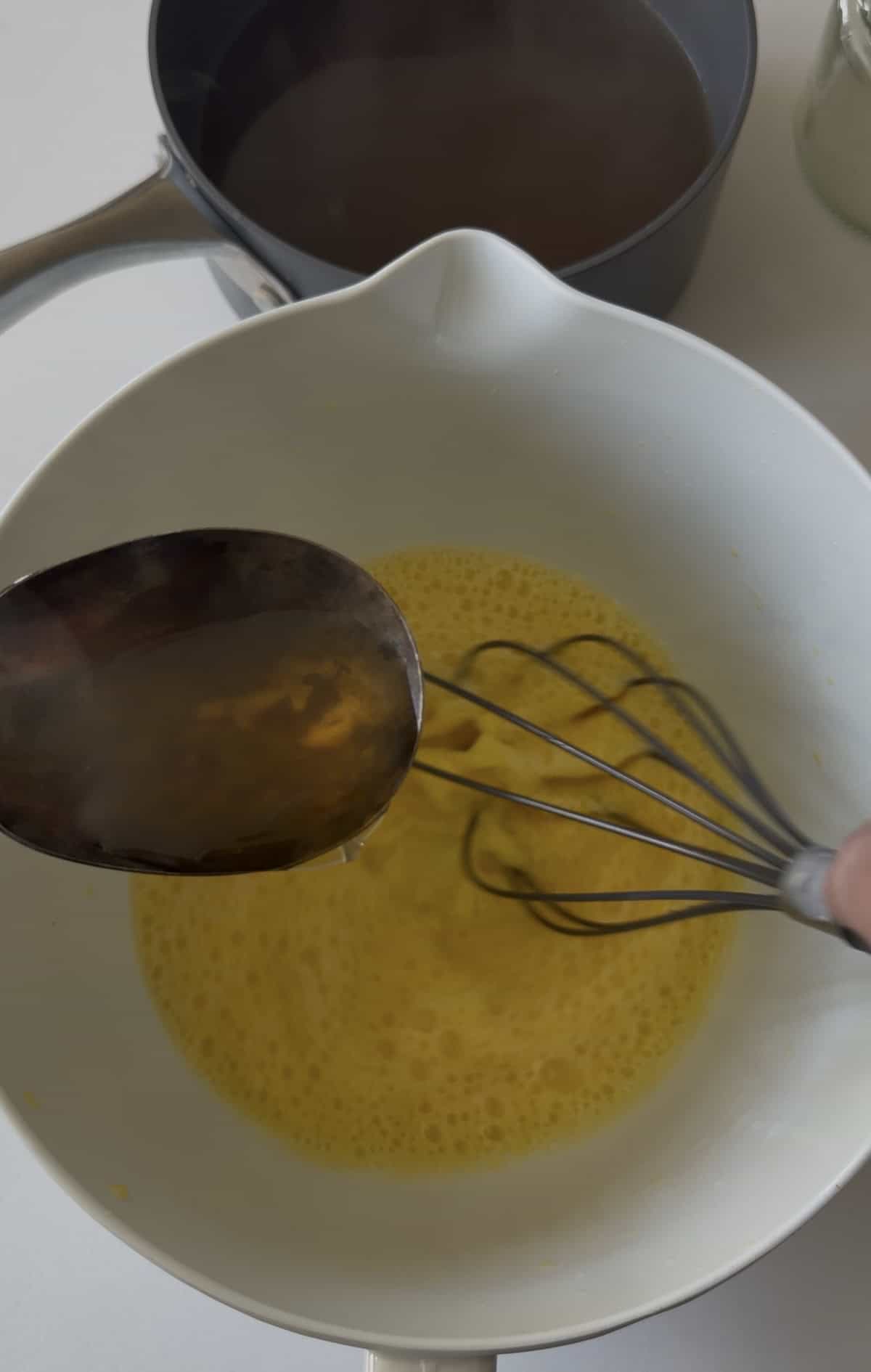 A metal spoon pours liquid into a bowl of beaten eggs being whisked, as Avgolemono sauce—a classic Greek sauce—comes together, with a saucepan and jar in the background.