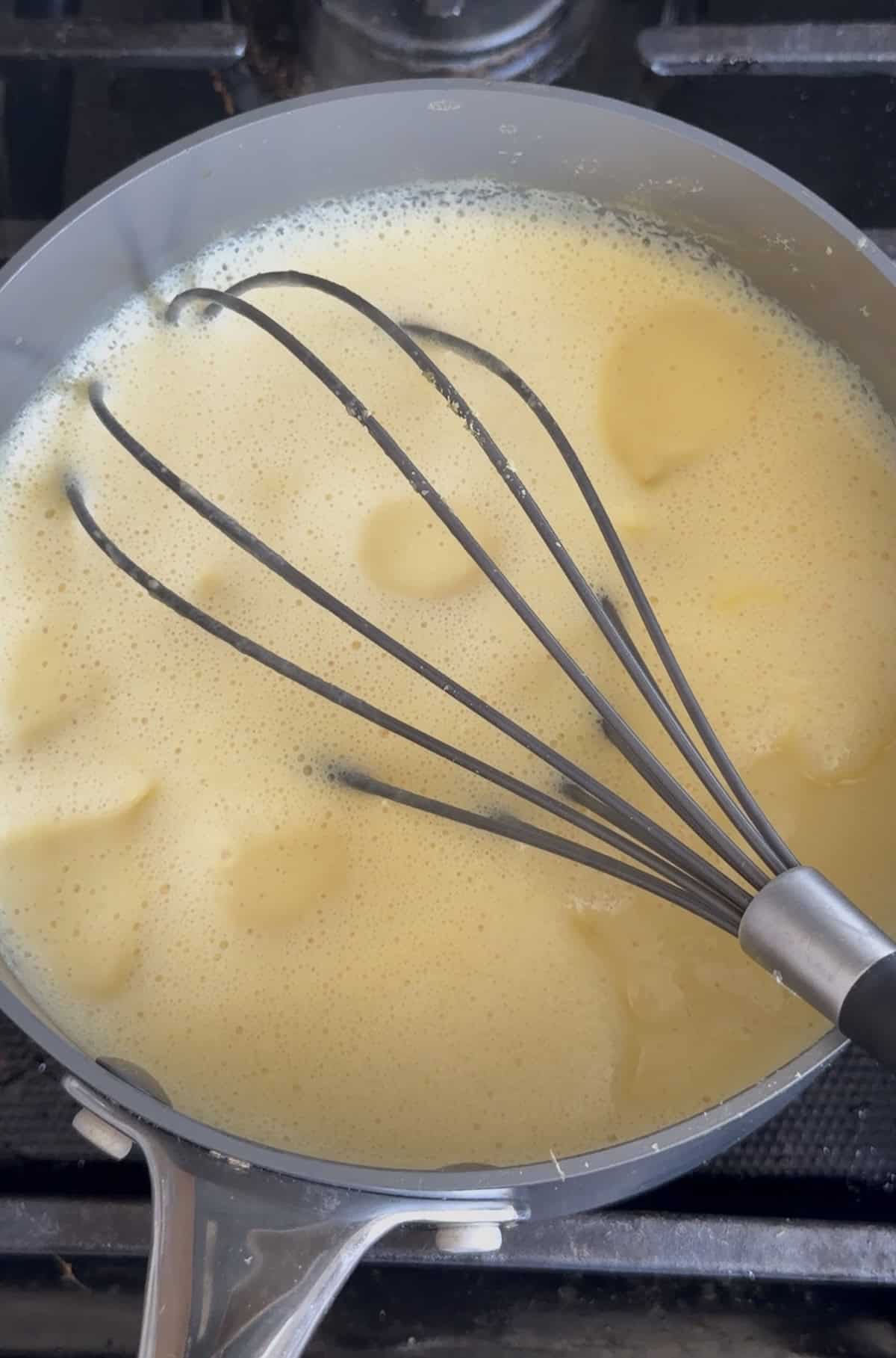 A metal whisk stirs a yellow avgolemono sauce, a classic Greek lemon egg sauce, in a saucepan on a stove.
