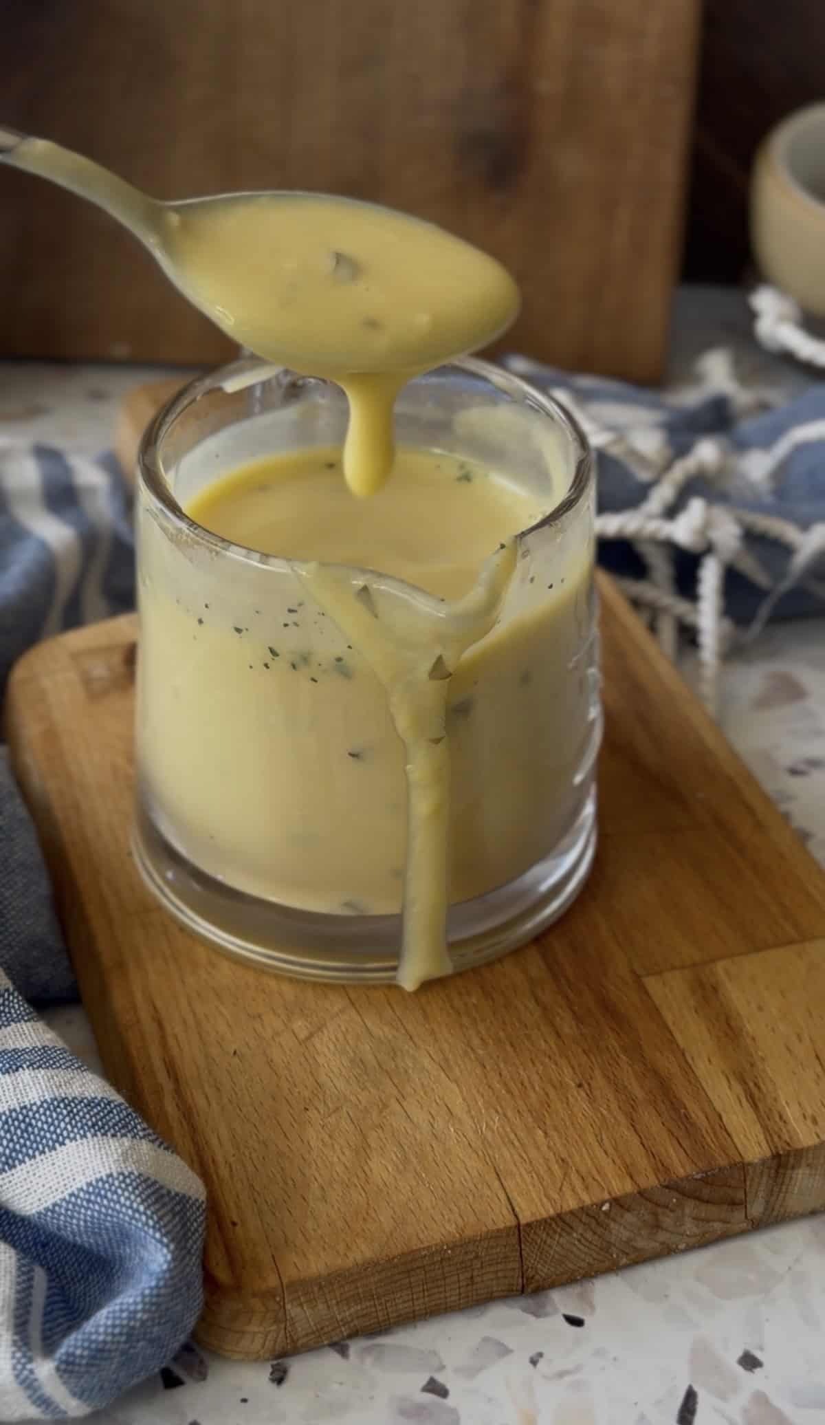 A spoon pours thick yellow Avgolemono Sauce with herbs into a glass cup, causing some sauce to drip down the side onto a wooden board.