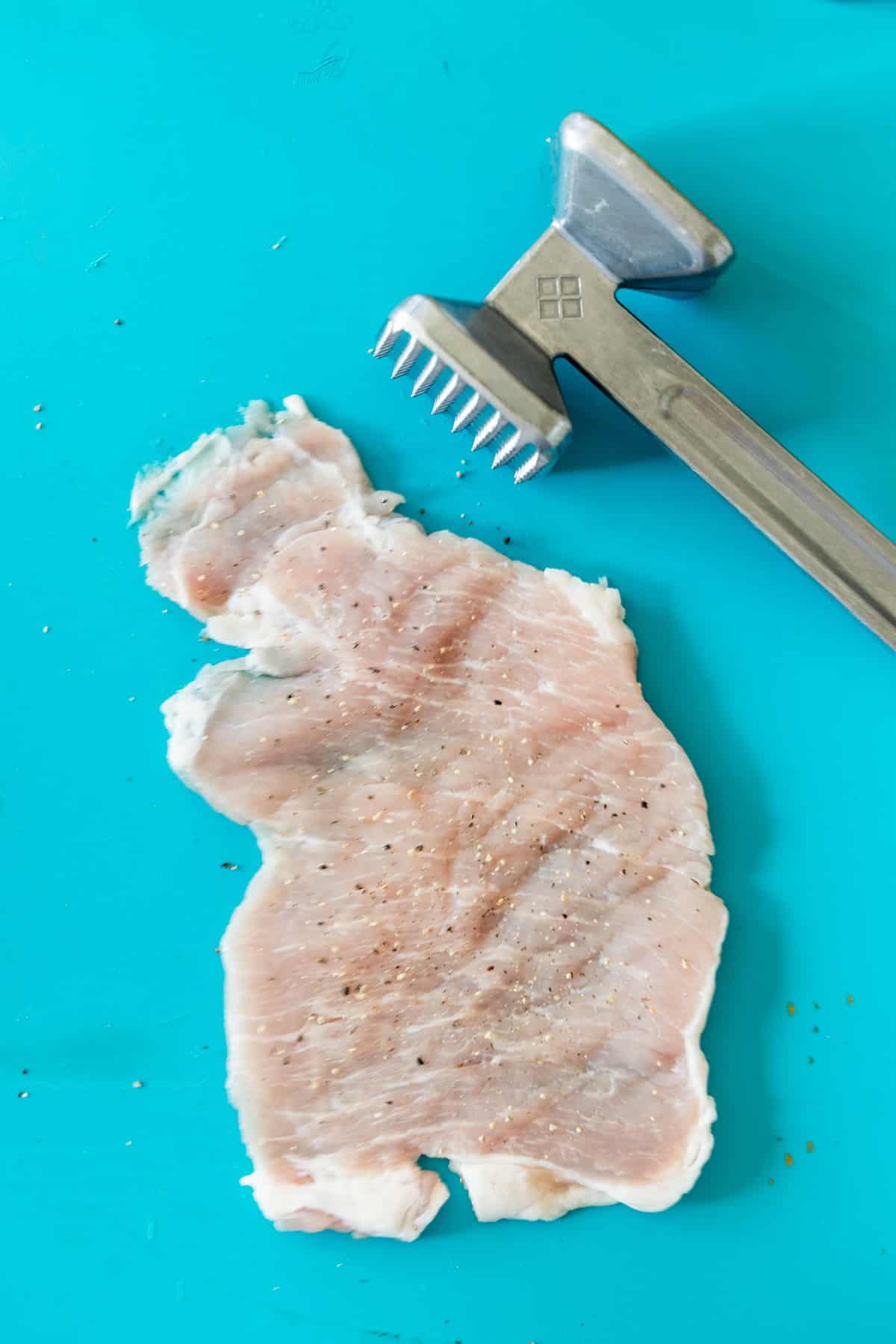 Raw chicken breast pounded flat on a blue surface next to a metal meat tenderizer, ready for recipes like Breaded Pork Chops or Baked Breaded Pork Chops.