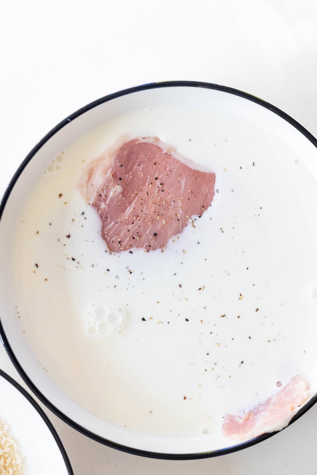 Raw meat slice soaking in a bowl of milk with black pepper sprinkled on top, preparing it for delicious baked breaded pork chops.