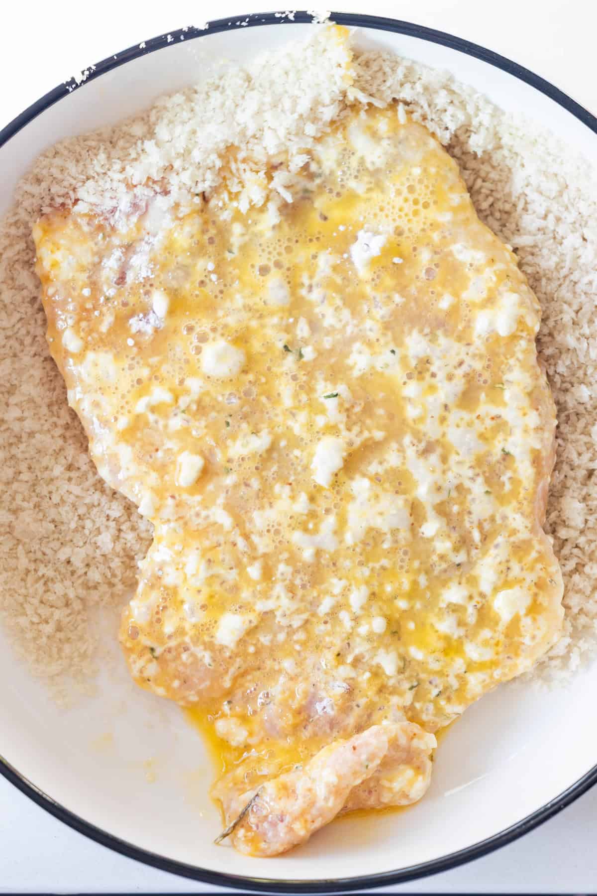 Raw chicken breast coated in beaten egg is placed on a layer of panko breadcrumbs in a white bowl, ready for breading&mdash;just like preparing Breaded Pork Chops or Baked Pork Chops.
