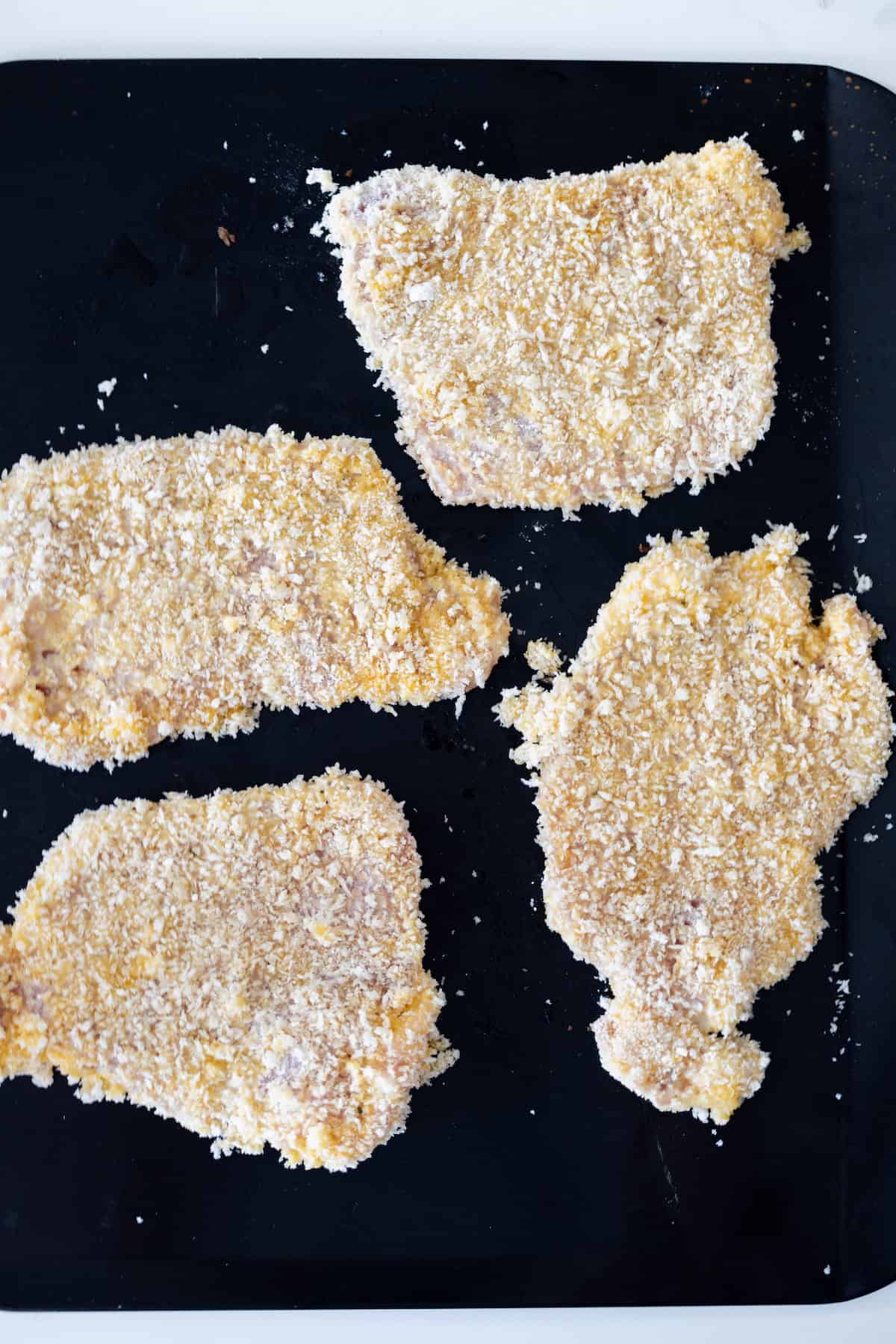 Four raw Breaded Pork Chops are arranged on a black surface, ready to be cooked into delicious Baked Breaded Pork Chops.
