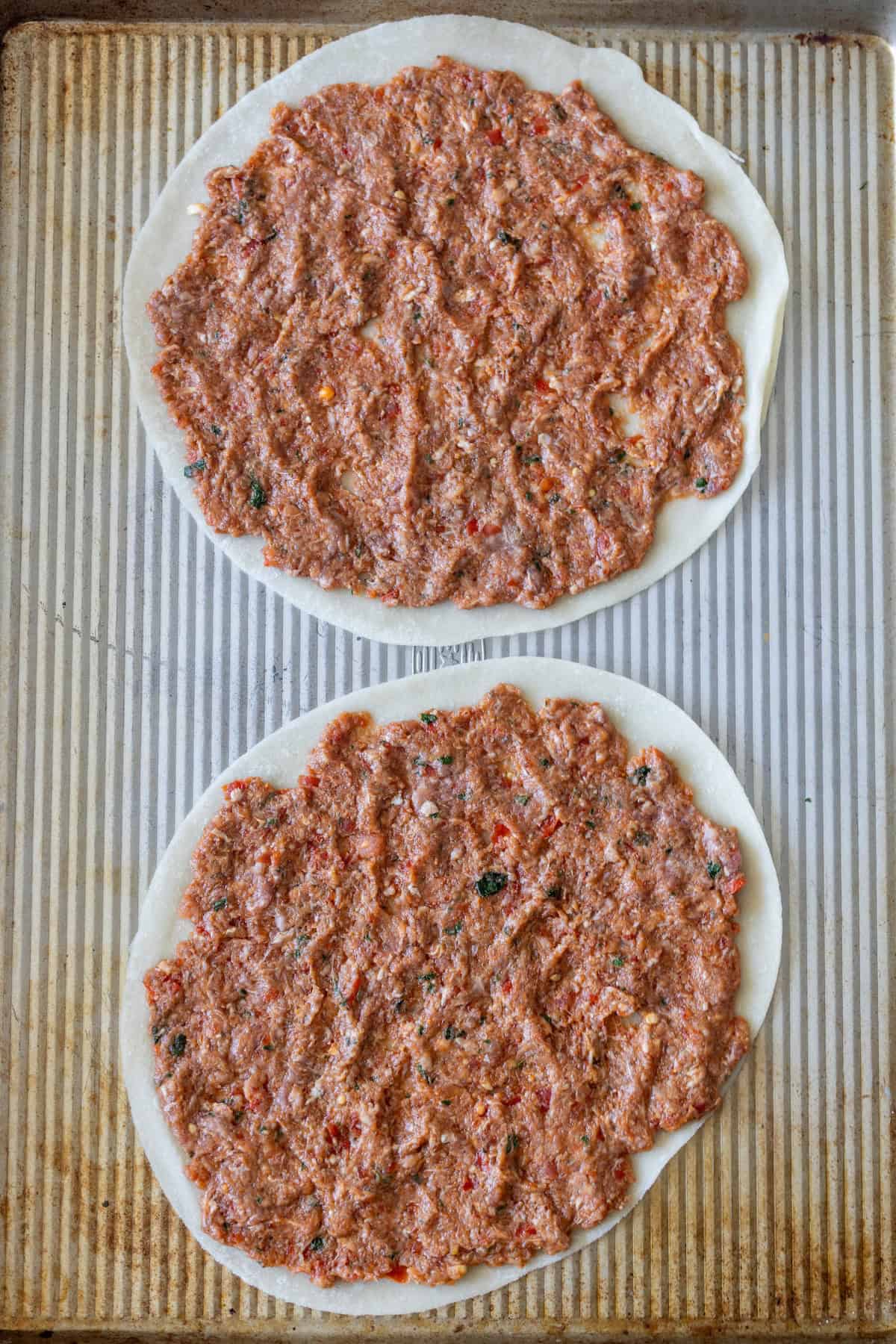 Two uncooked Armenian pizza flatbreads, known as Lahmajoun, rest on a baking sheet with seasoned ground meat spread evenly—an easy recipe for a delicious meal.