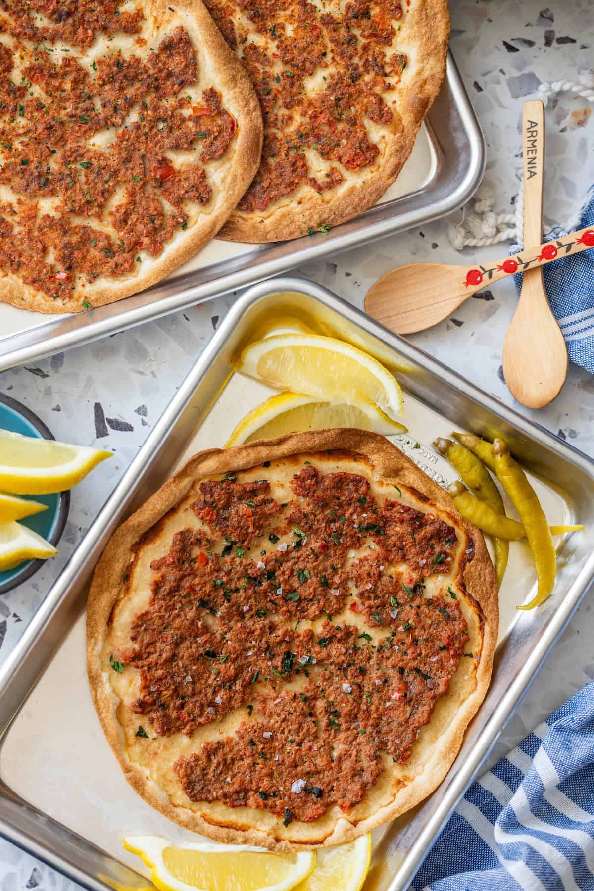 A tray of Easy Lahmajoun, Armenian flatbreads topped with minced meat and herbs, is served with lemon wedges and green chili peppers. Another tray of these delicious flatbreads can be seen in the background.