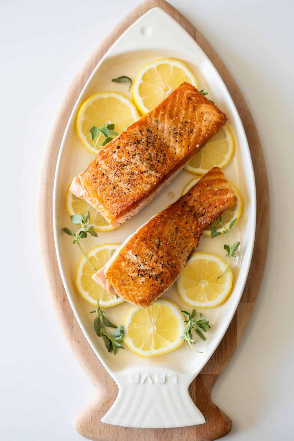 Two pan fried salmon filets served on a fish-shaped plate, garnished with lemon slices and fresh herbs.