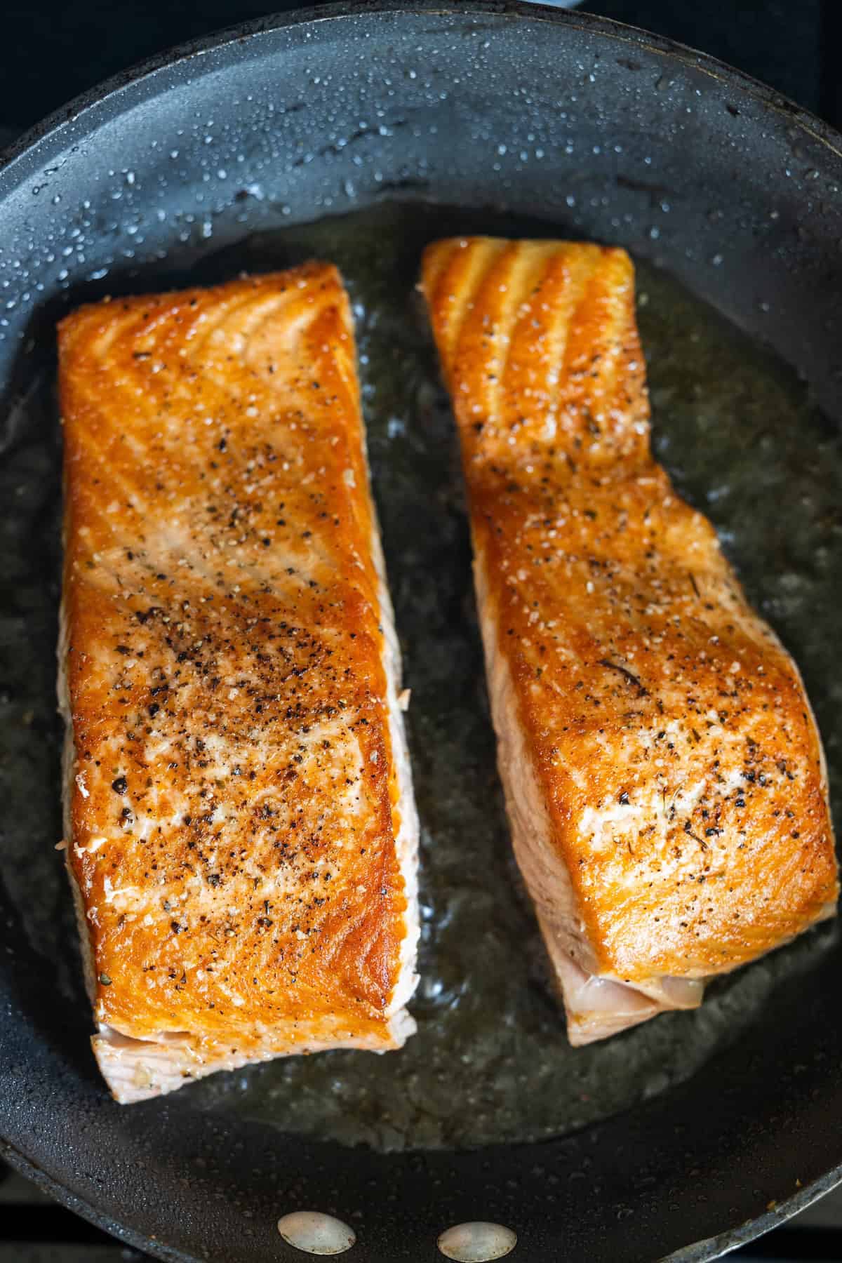 Two seasoned salmon filets being pan-seared to perfection in a black skillet with oil, creating delicious pan fried salmon.