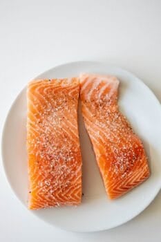 Two raw salmon filets with visible marbling, seasoned with salt and pepper, arranged side by side on a white plate&mdash;perfect for making delicious pan fried salmon.