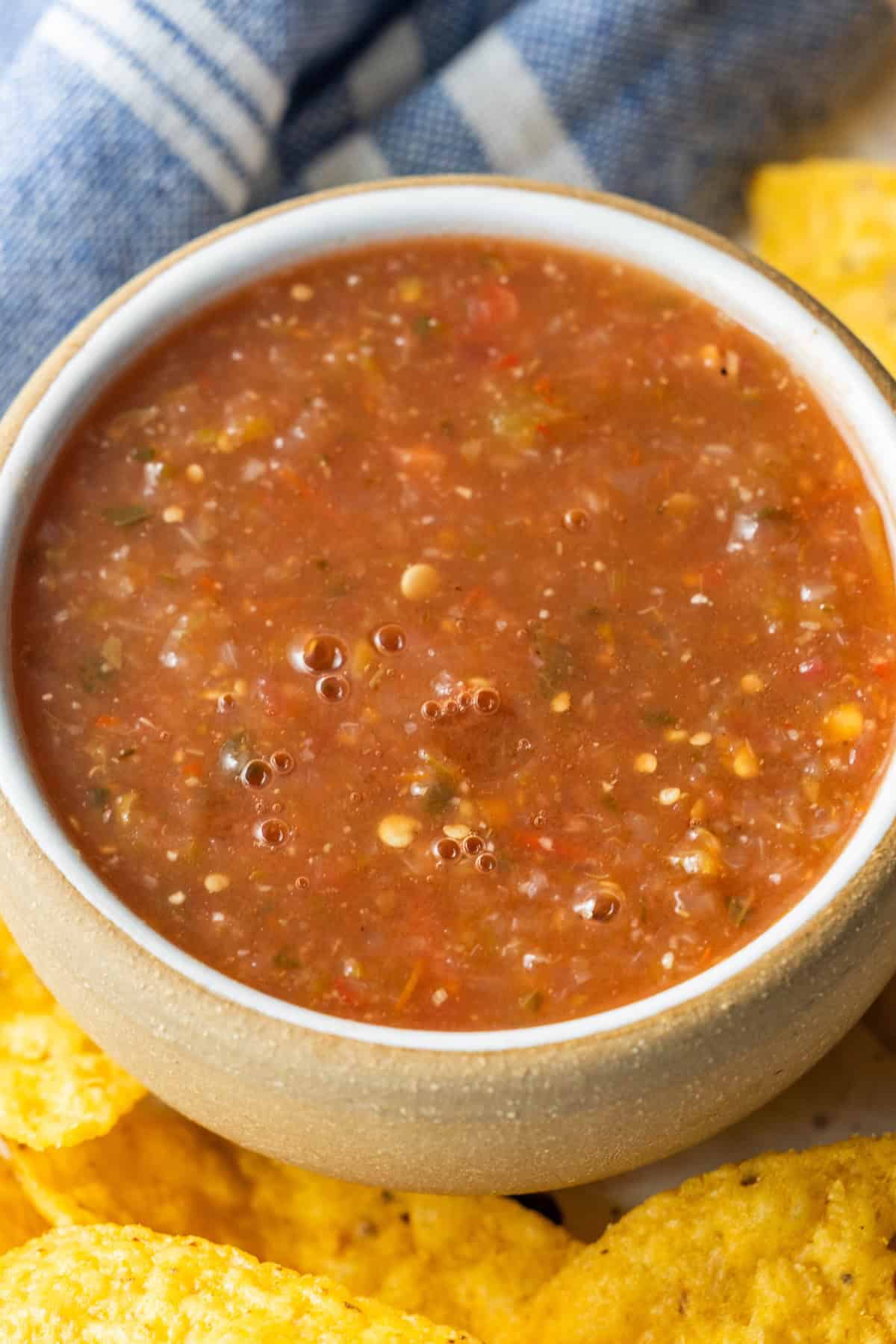 A bowl of chunky roasted salsa sits next to yellow tortilla chips with a blue and white cloth in the background.