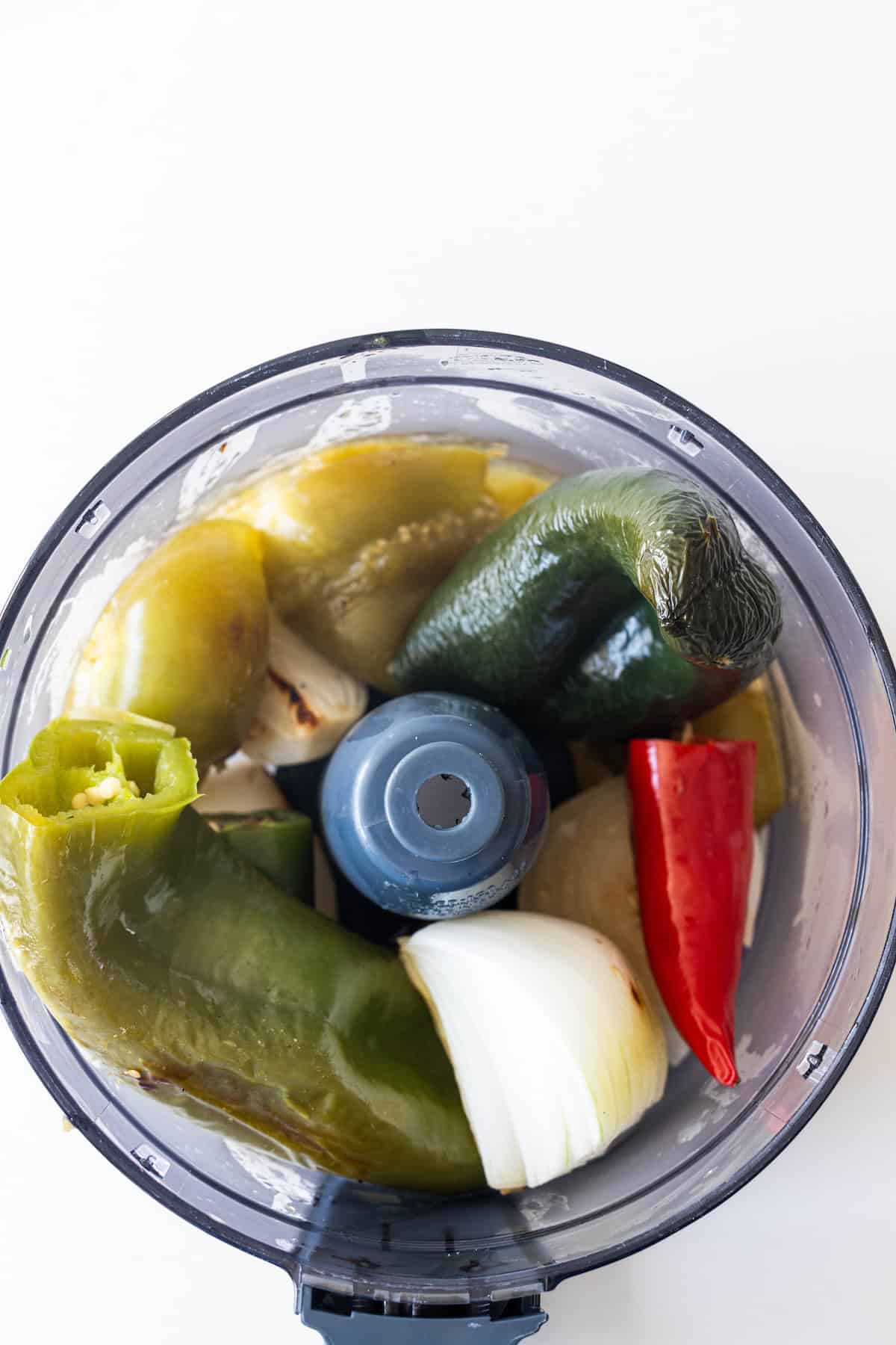 A food processor bowl containing chunks of roasted green and red peppers, pieces of onion, and the makings of a vibrant roasted salsa, viewed from above.