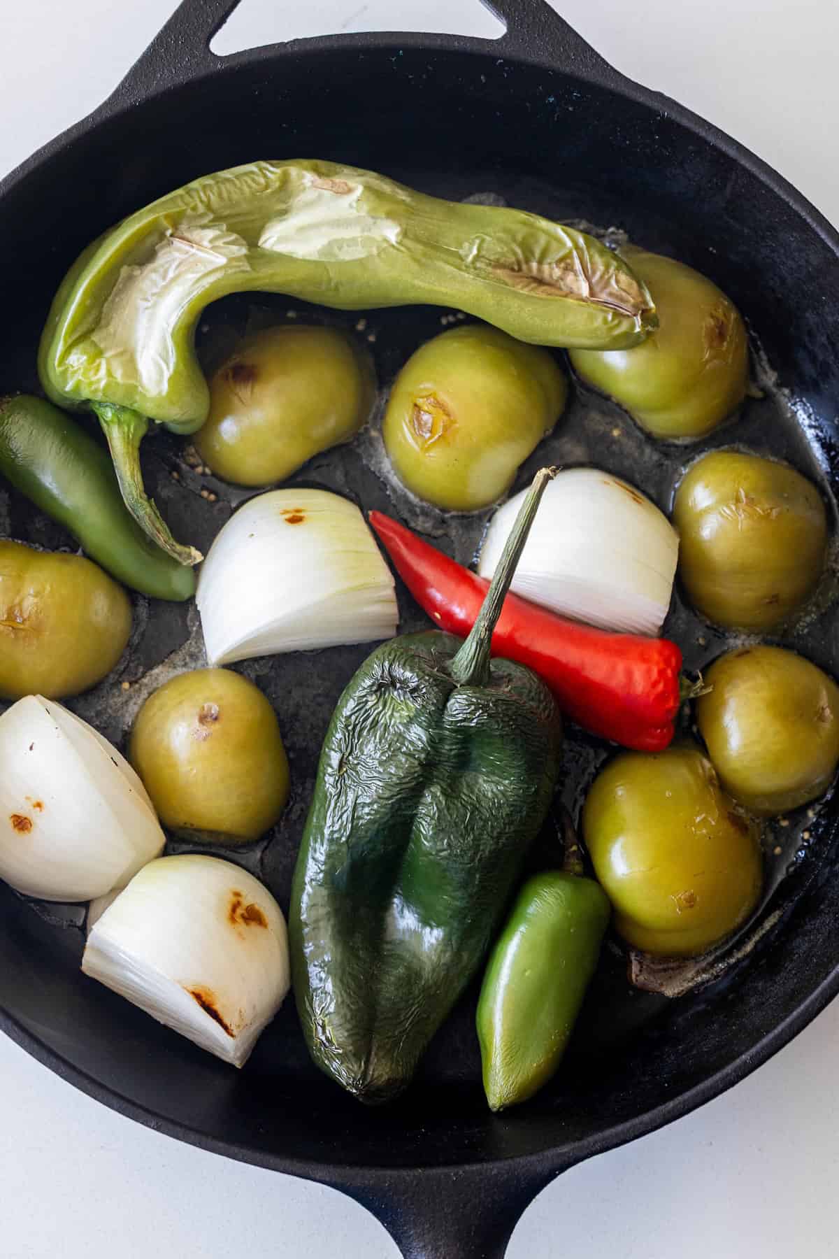 A cast iron skillet with tomatillos, white onion chunks, jalape&ntilde;o, poblano, Anaheim, and red chili peppers being roasted for a flavorful roasted salsa.