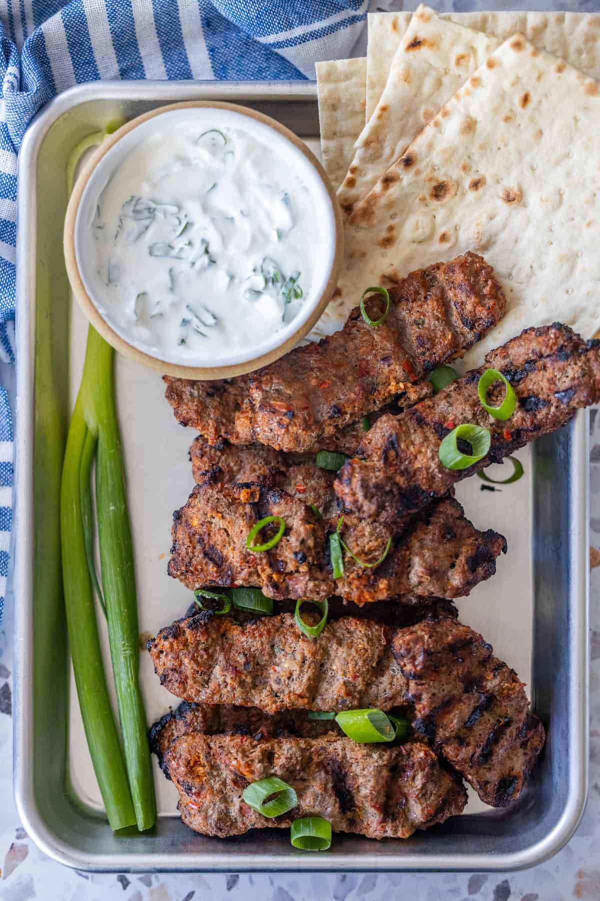 A metal tray with Armenian spiced meat kebabs, sliced green onions, flatbread, two whole green onions, and a bowl of yogurt sauce.