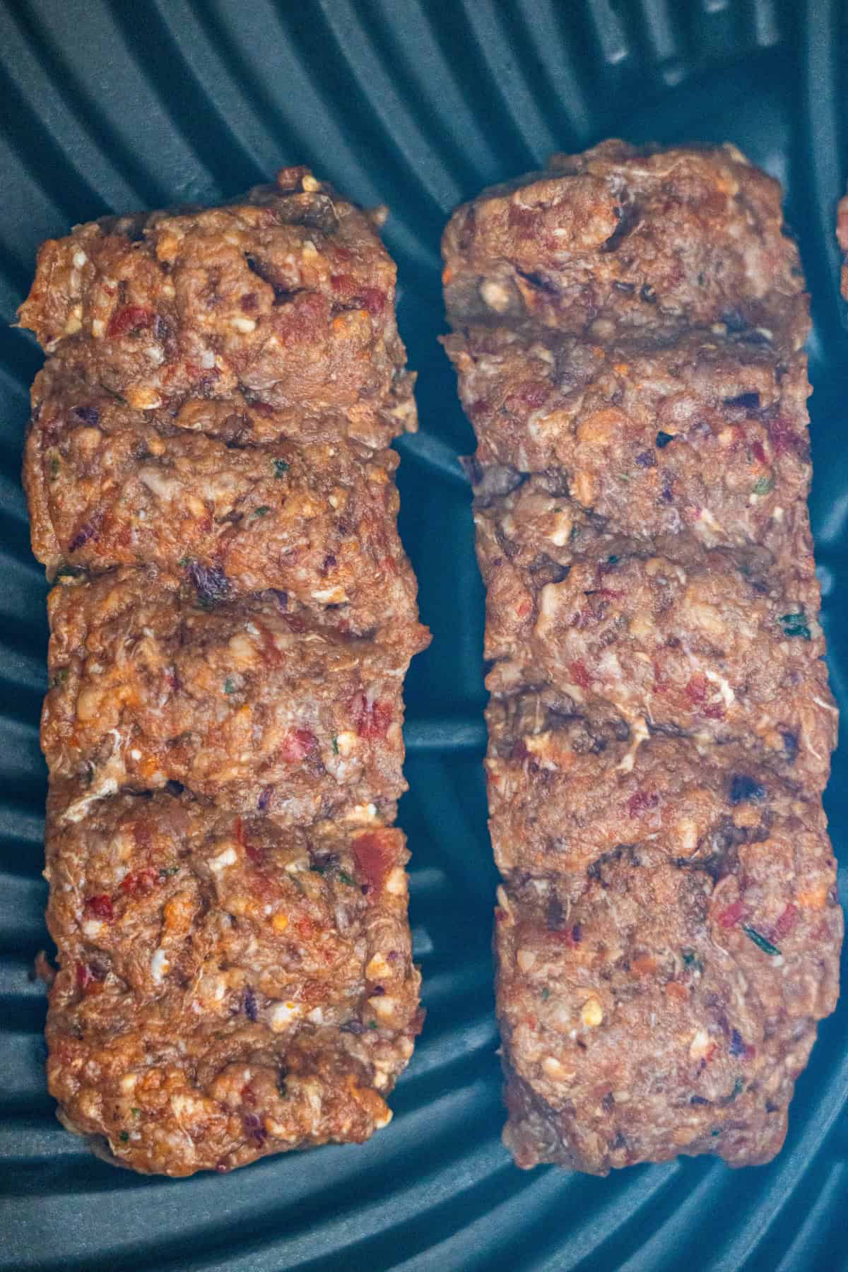 Two browned Armenian kebabs made of spiced meat with visible herbs and spices are cooking on a black ridged grill surface.