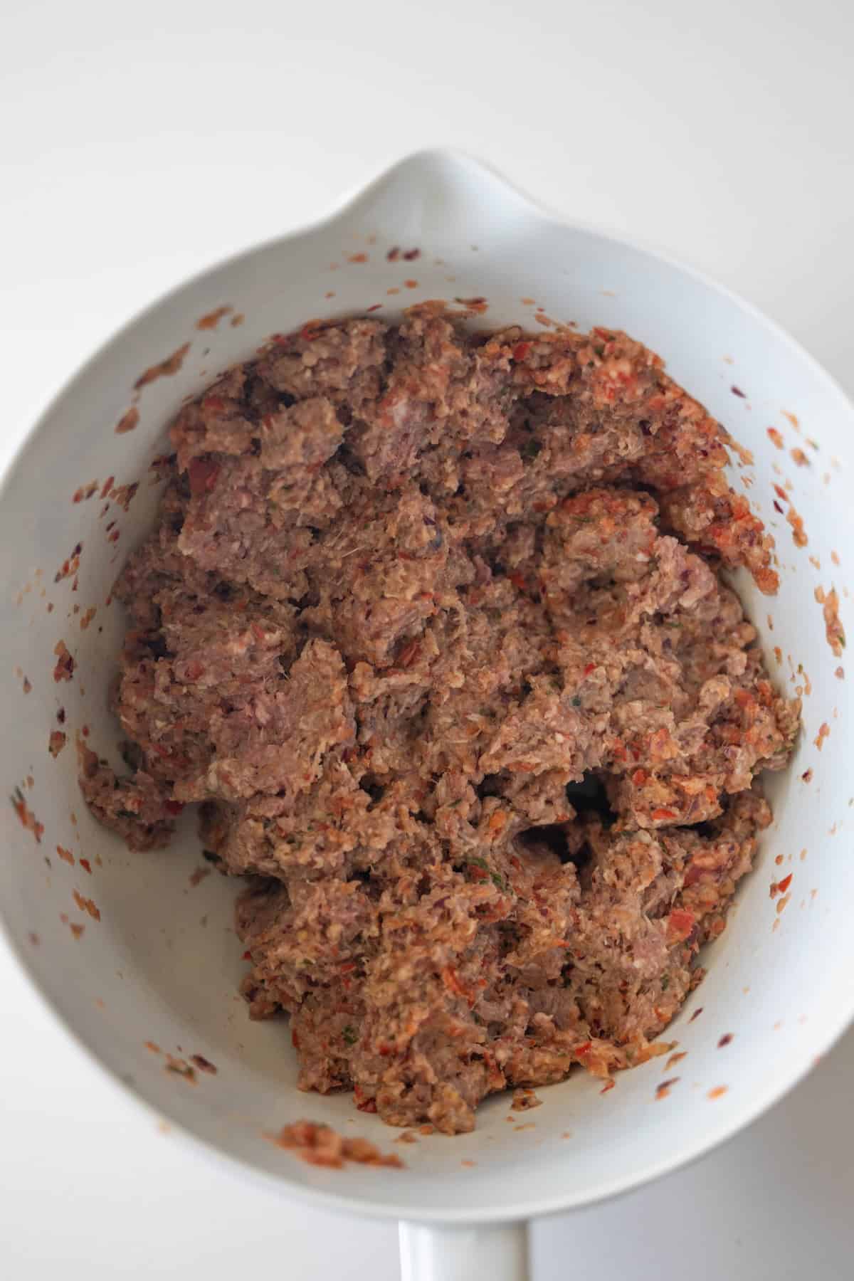 A white mixing bowl containing Armenian spiced meat and chopped vegetables, perfect for making flavorful kebabs, sits on a white surface.