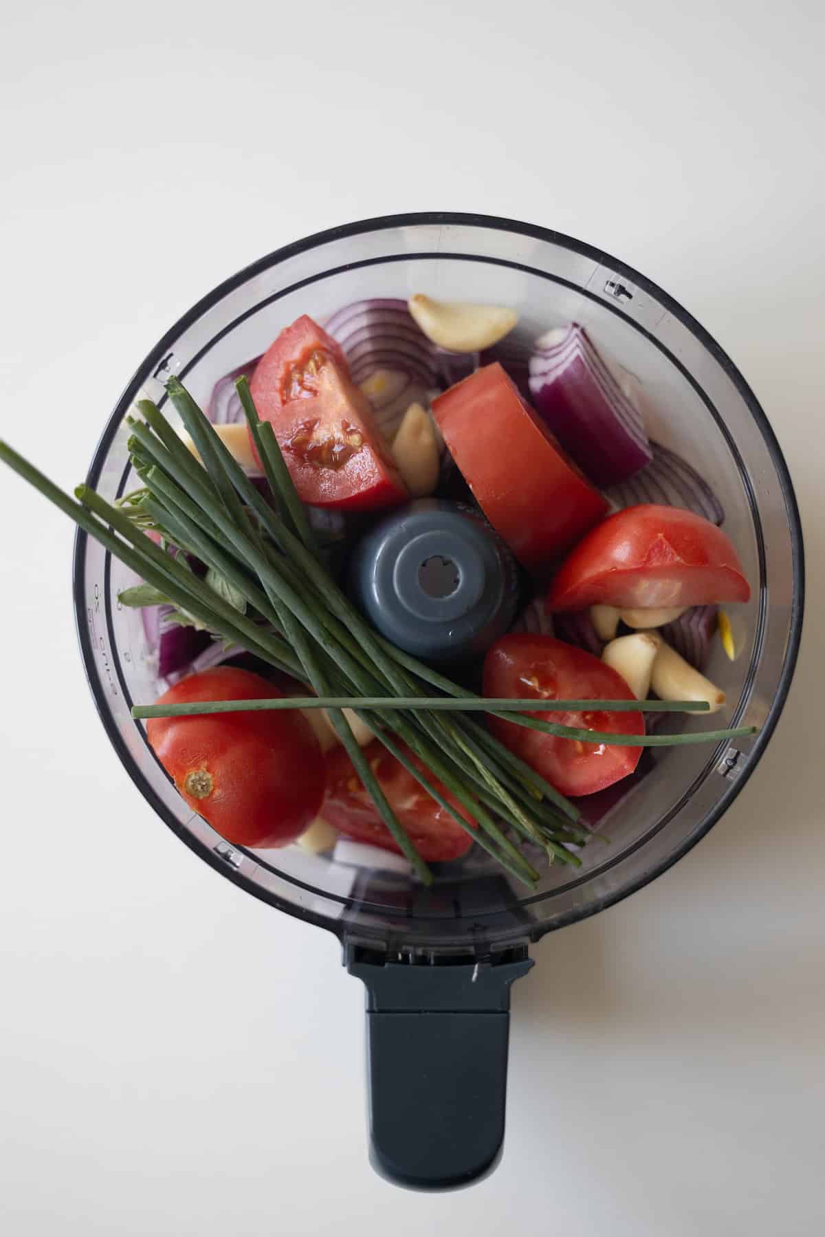 A food processor bowl filled with chopped red onion, tomatoes, garlic cloves, and fresh chives—perfect for preparing Armenian kebabs or spiced meat mixtures, viewed from above.