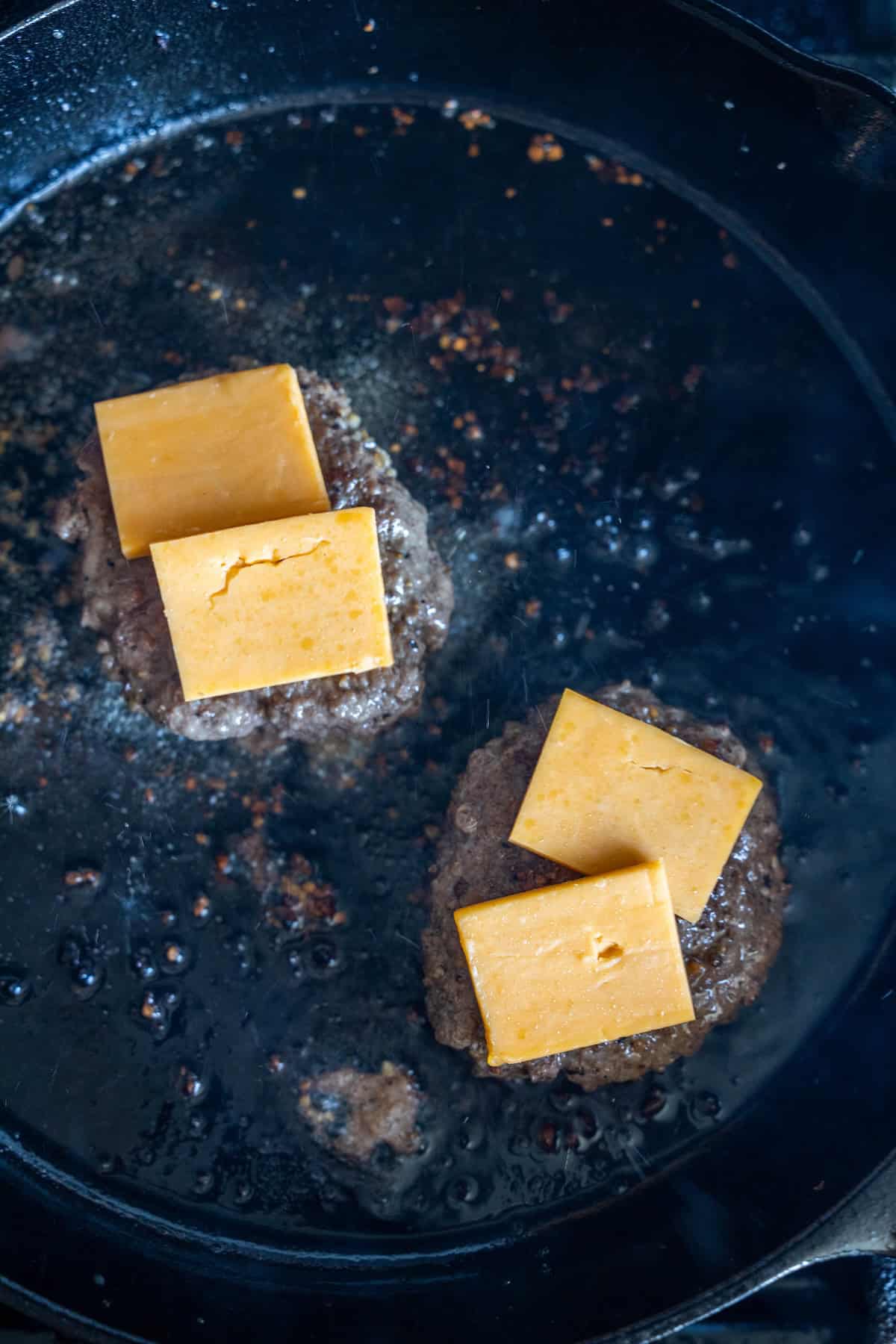 Two burger patties sizzle in a cast iron skillet as cheddar cheese melts on top—perfect for low carb meals or creative cheeseburger recipes.