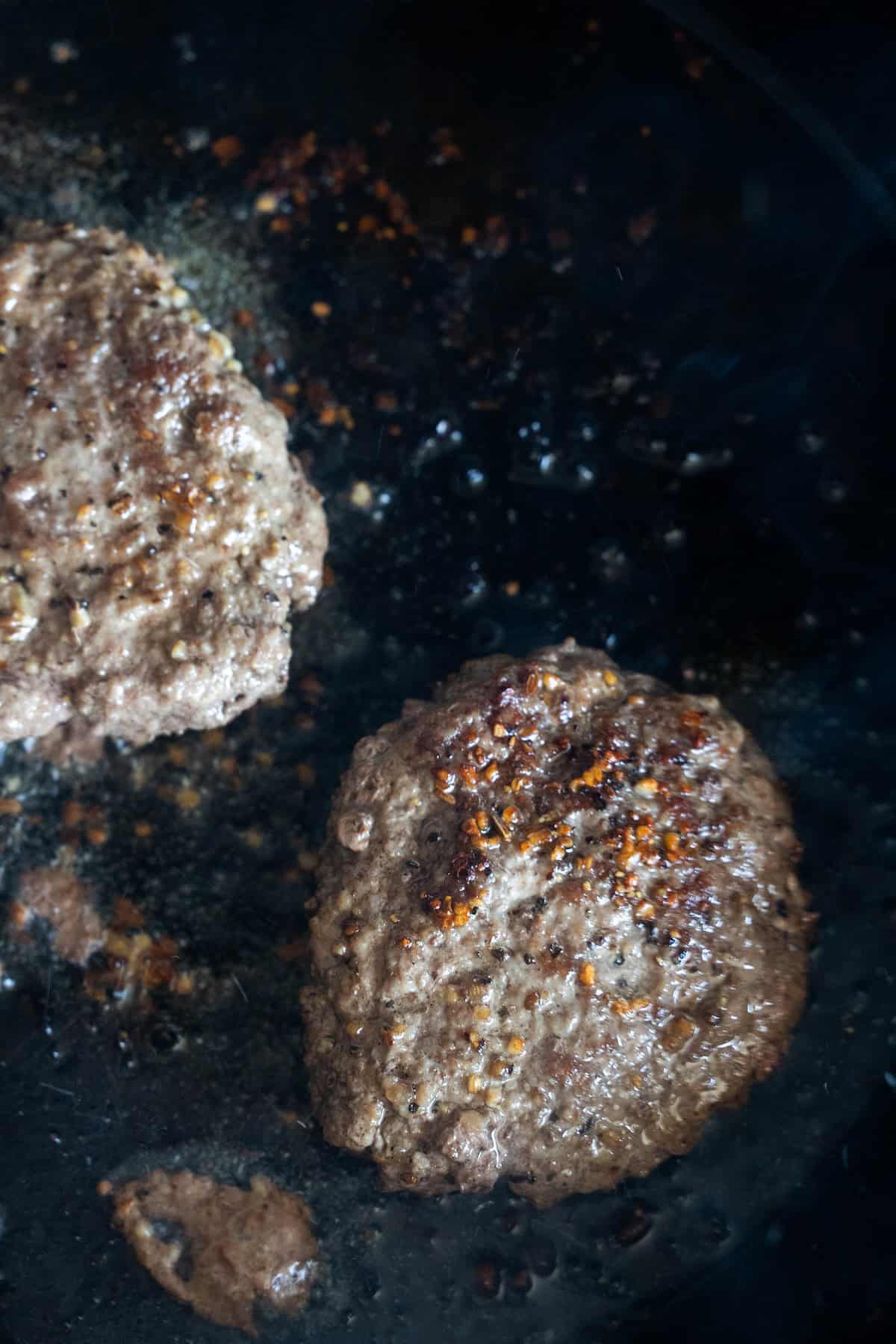 Two beef burger patties cooking in a dark pan, with visible oil and seasoning on the surface—perfect for creating a flavorful burger bowl or trying out your favorite cheeseburger recipe.