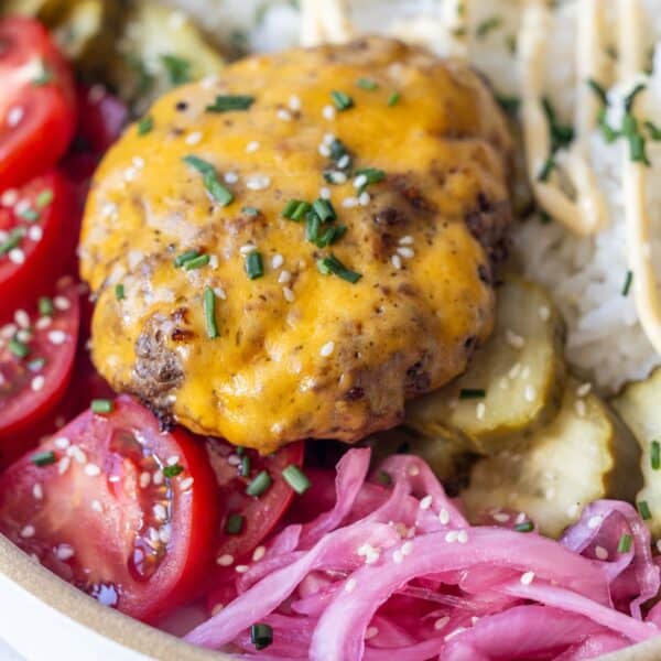 A close-up of a Cheeseburger Bowl features a cheddar-topped beef patty, sliced tomatoes, pickles, pickled onions, rice, sesame seeds, chives, and a drizzle of sauce—a fresh twist on the classic cheeseburger recipe.