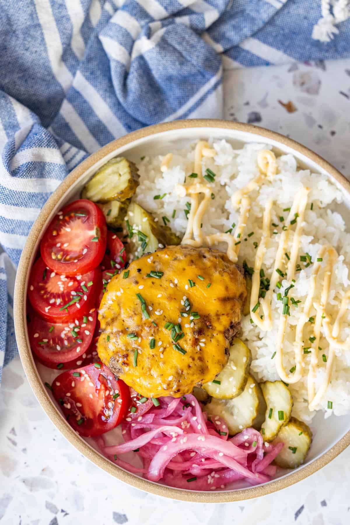 A bowl of white rice topped with a cheddar-topped burger patty, sauce, sliced pickles, cherry tomatoes, and pickled red onions, garnished with chives—a delicious cheeseburger bowl inspired by your favorite cheeseburger recipe.